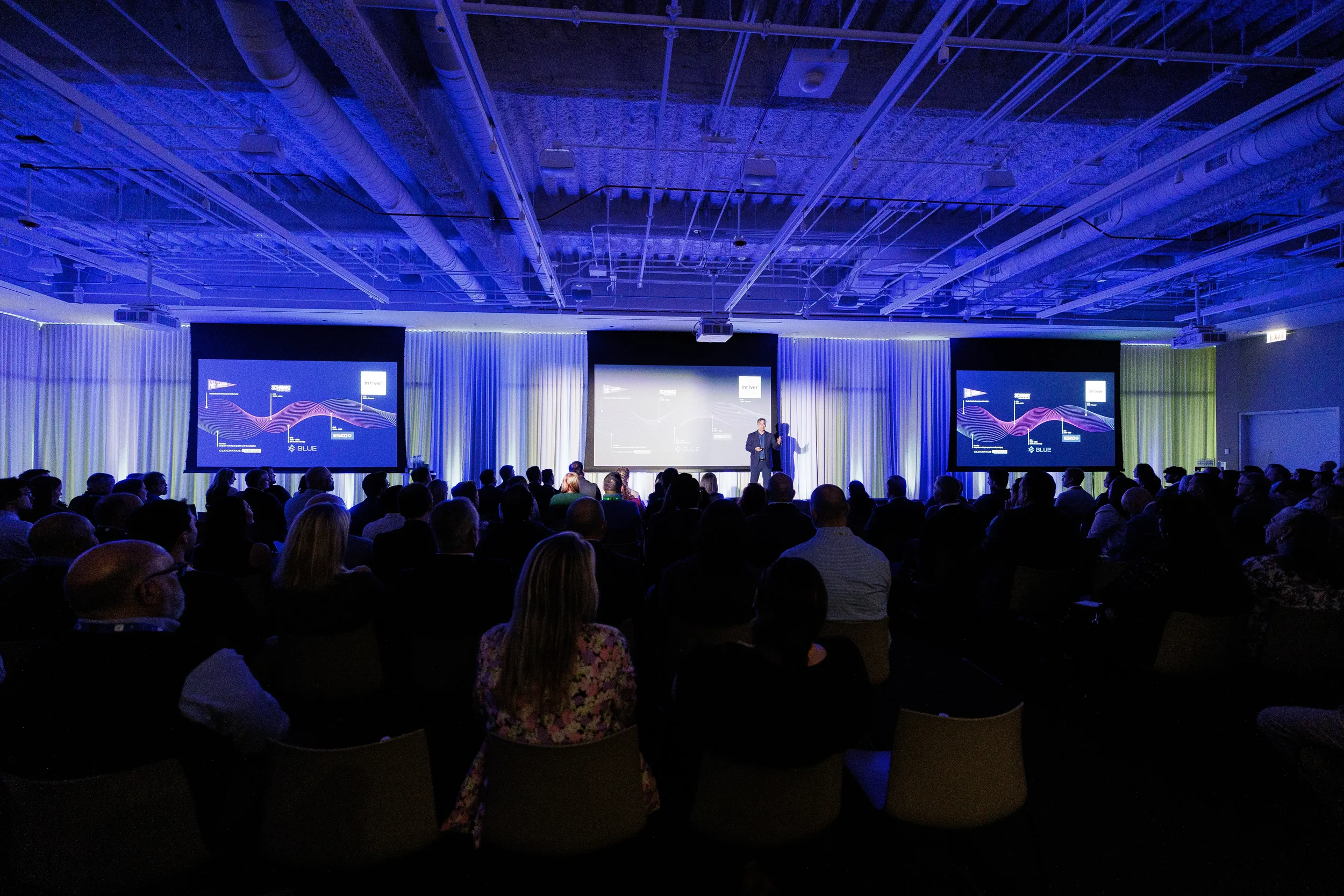 Audience watches speaker on stage before three projection screens in blue-lit Chicago corporate conference