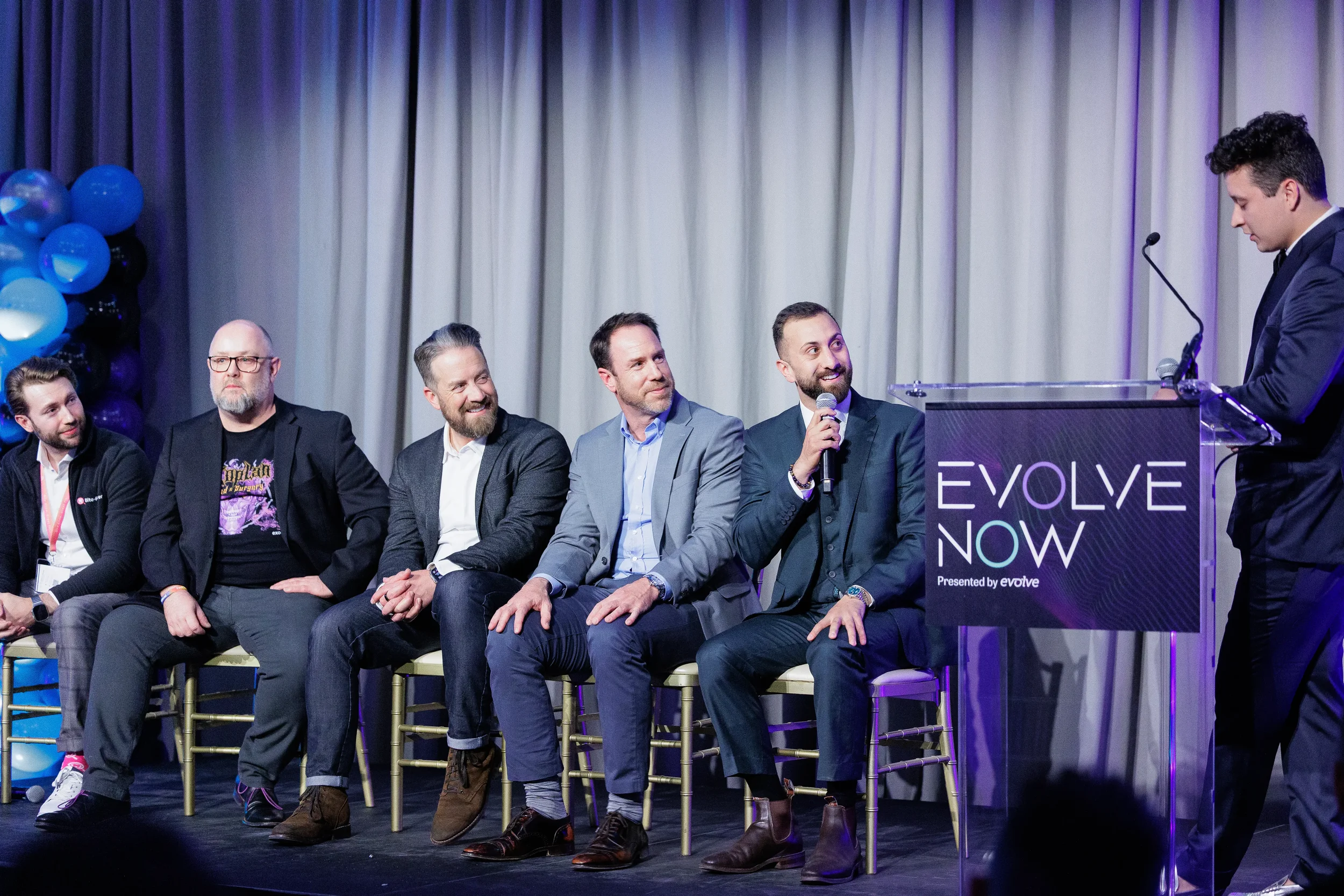 Five panelists seated on stage beside branded podium with balloon décor at Chicago corporate event