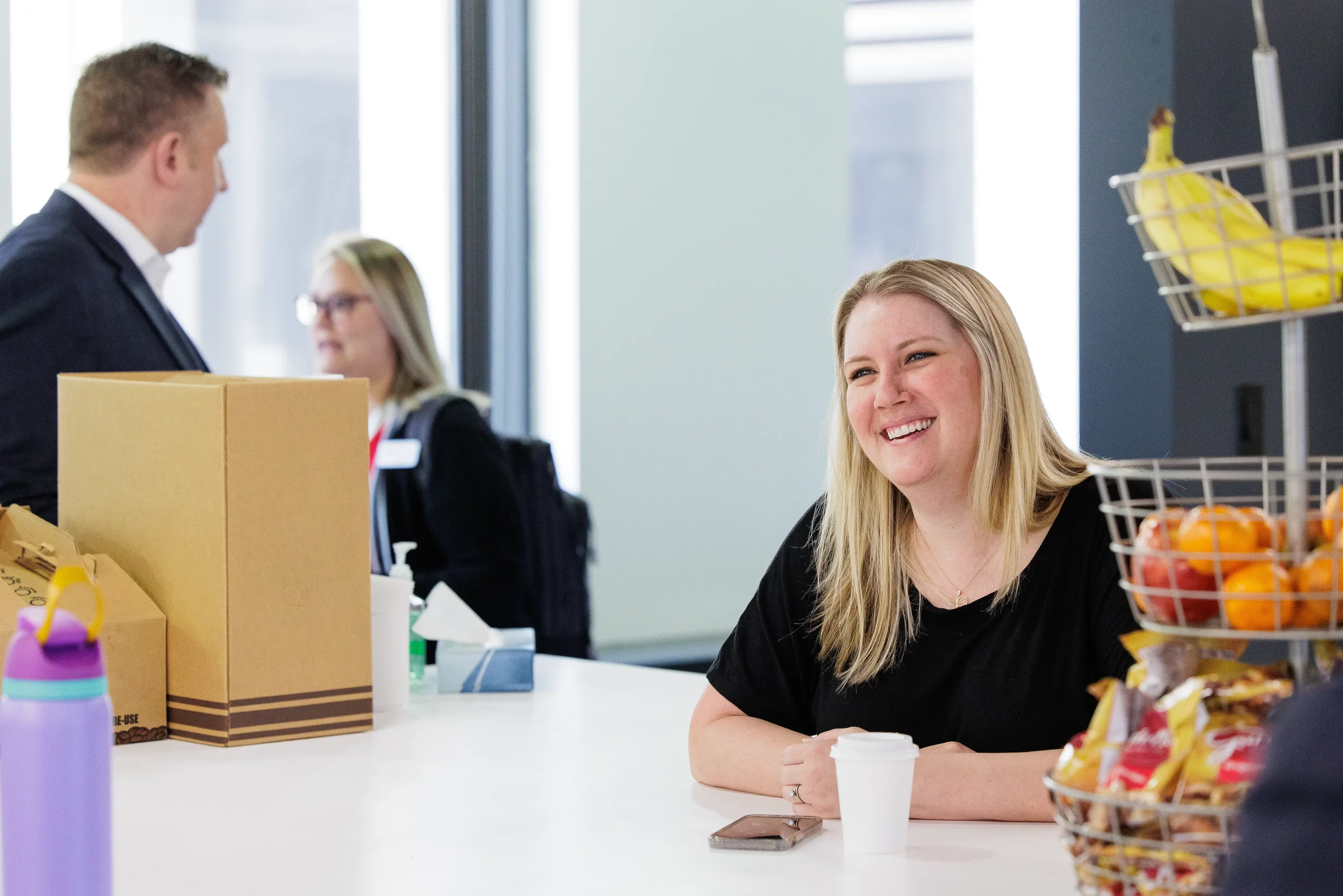 Blond woman in a black shirt sits at a table near a fruit basket, smiling, with others blurred in the background in Chicago