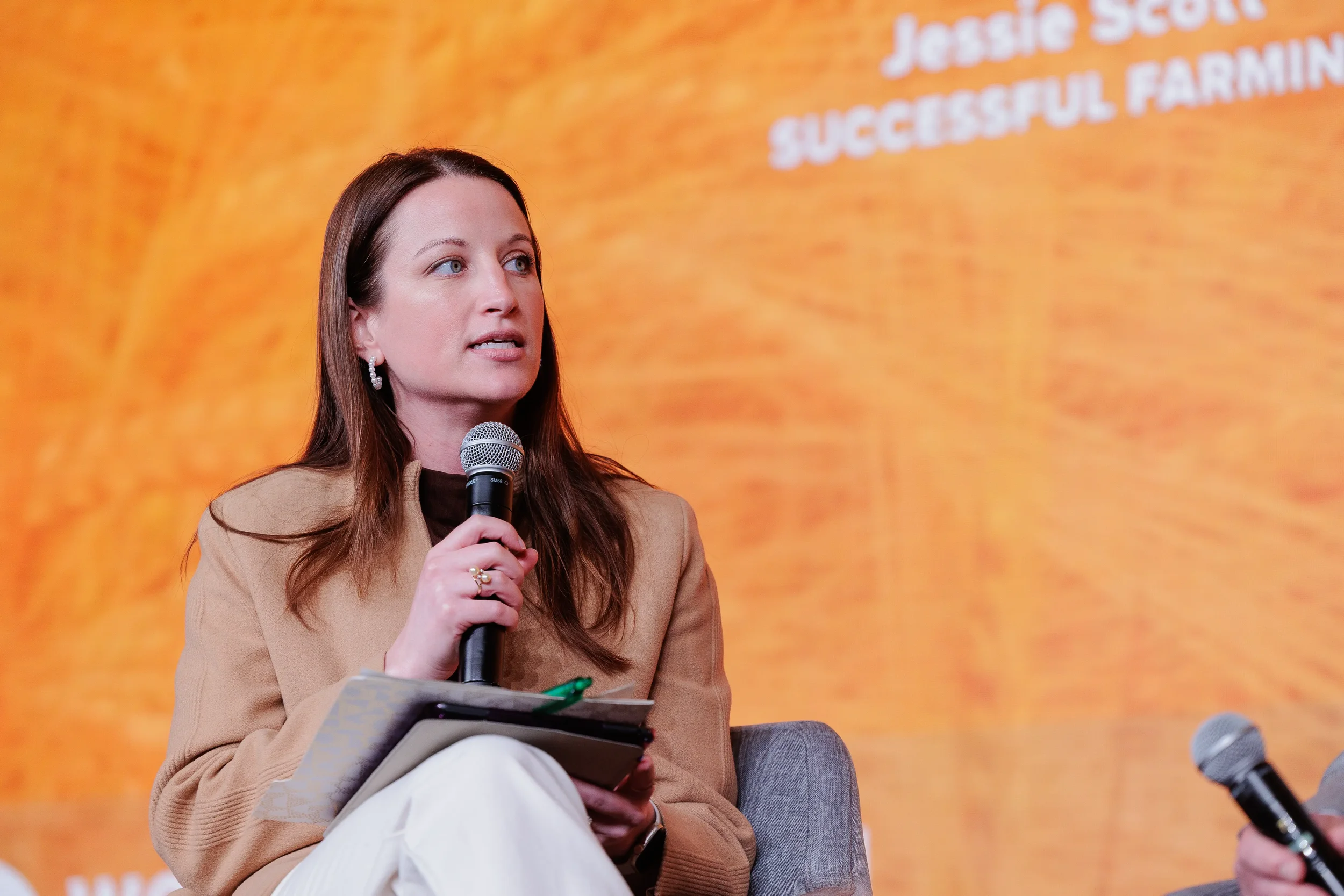 Female panelist in tan blazer holds microphone on stage before orange backdrop at Chicago industry conference