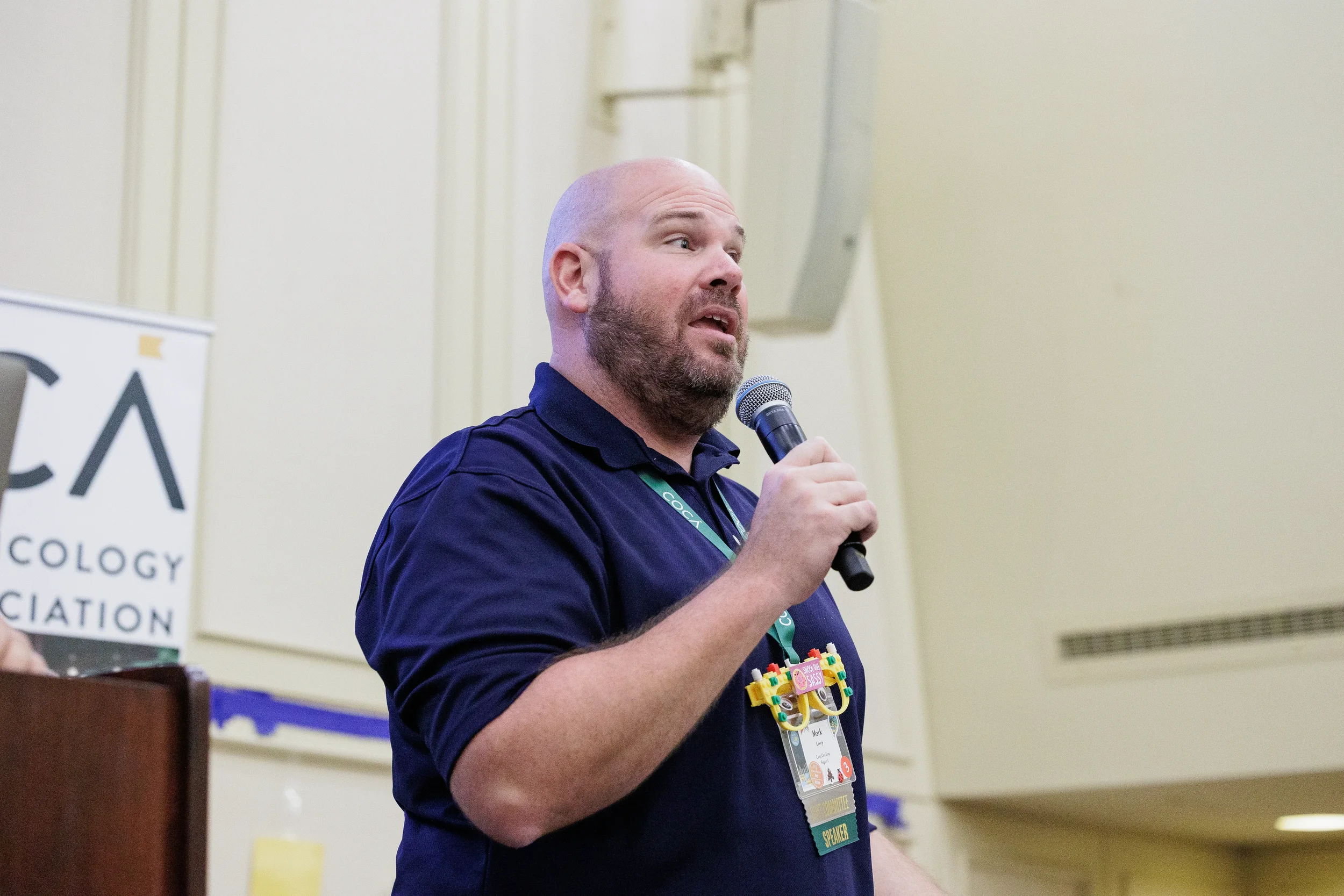 Speaker holds microphone and addresses audience beside branded COCA signage at Orlando nonprofit conference event