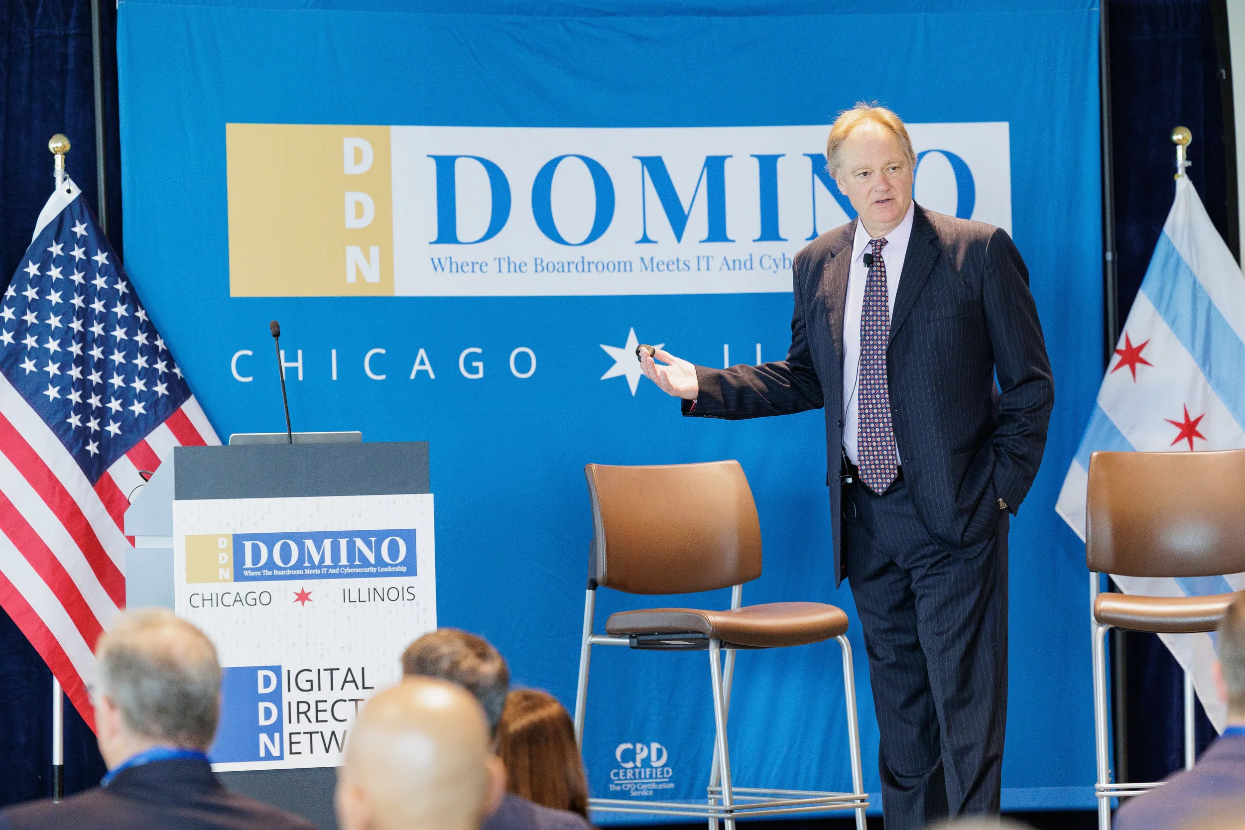 Male speaker gestures beside branded podium on stage with flags at Orlando corporate conference event