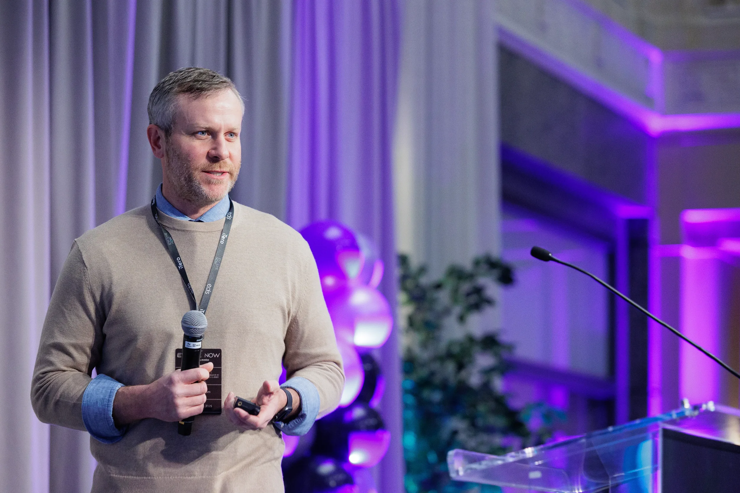 Lanyard-wearing speaker holds microphone near podium with purple event backdrop at Chicago industry conference