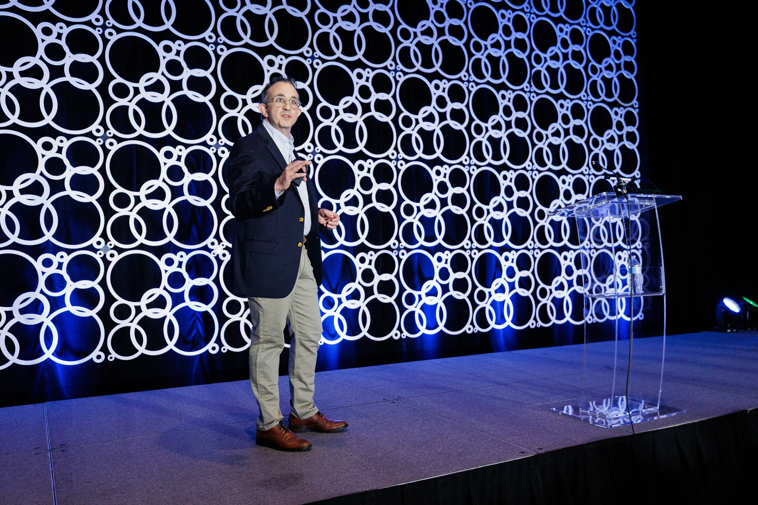 Presenter speaks on stage with circle-patterned backdrop beside clear podium at Orlando industry conference