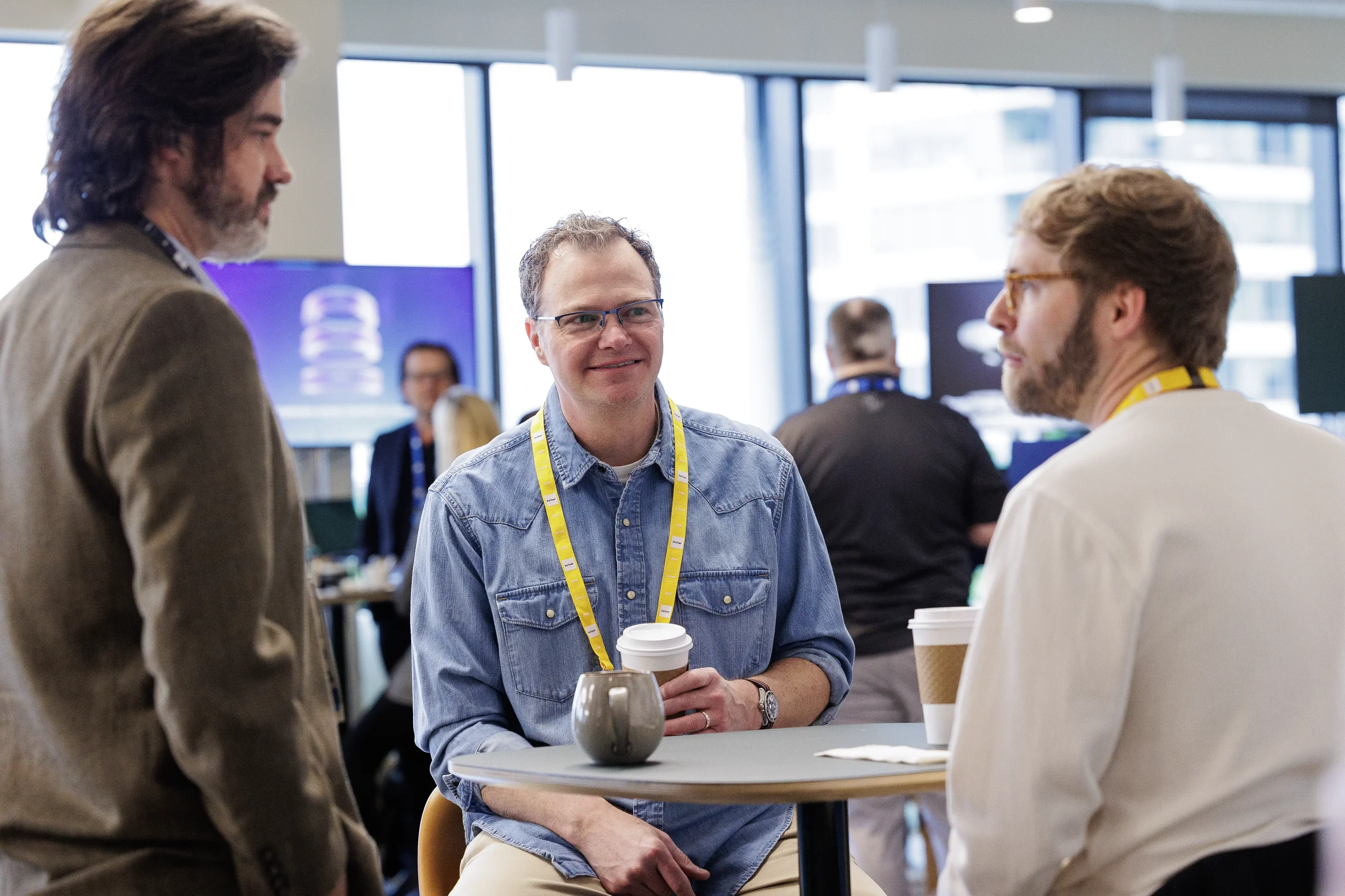Smiling attendees with lanyards chat over coffee at table in lounge during Orlando corporate conference