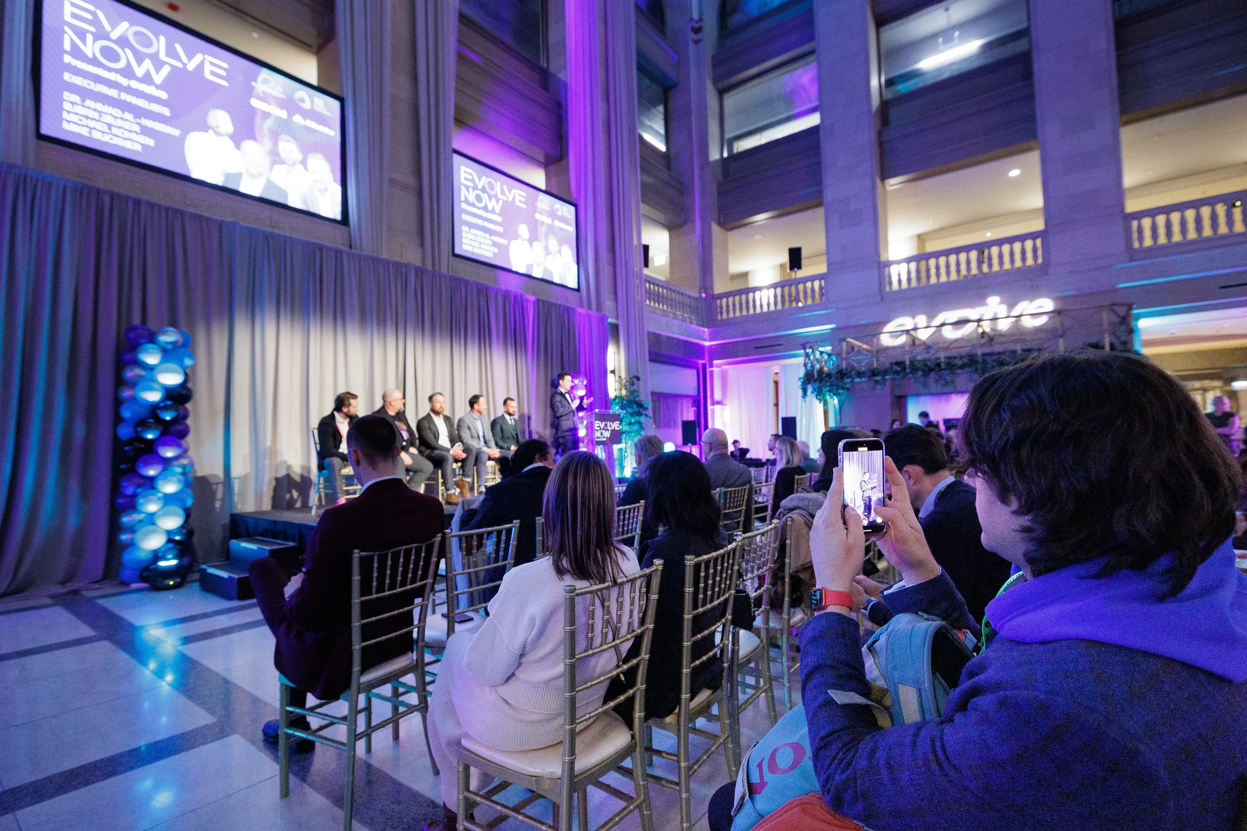 Attendee photographs panel discussion on phone during Chicago industry conference in purple-lit atrium