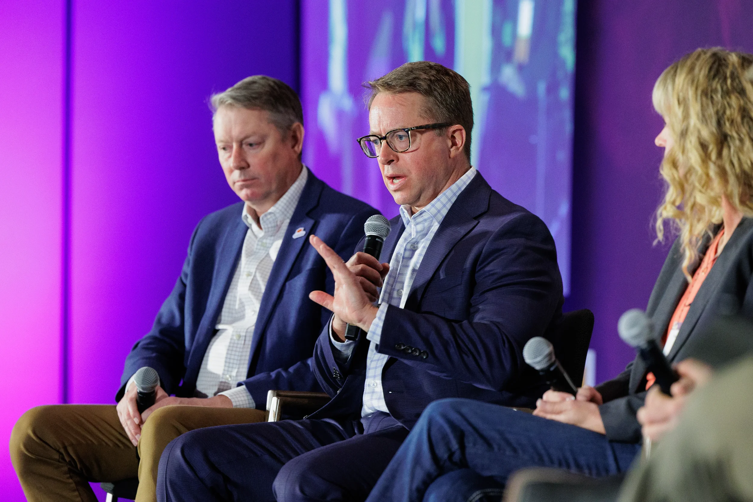 Panelist gestures while speaking into microphone during seated discussion at Orlando corporate summit