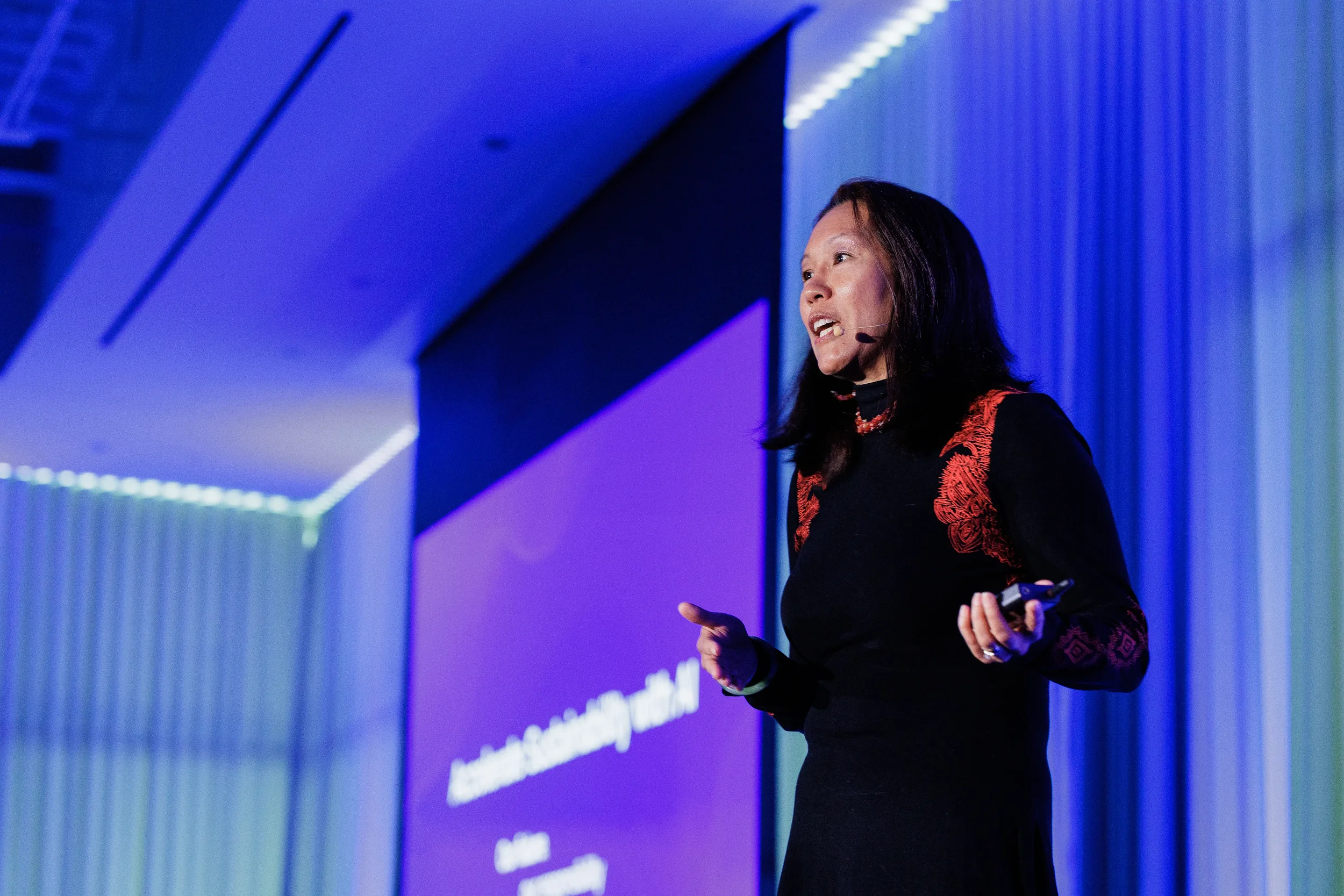Speaker with headset microphone gestures while presenting beside purple slide at Orlando industry conference