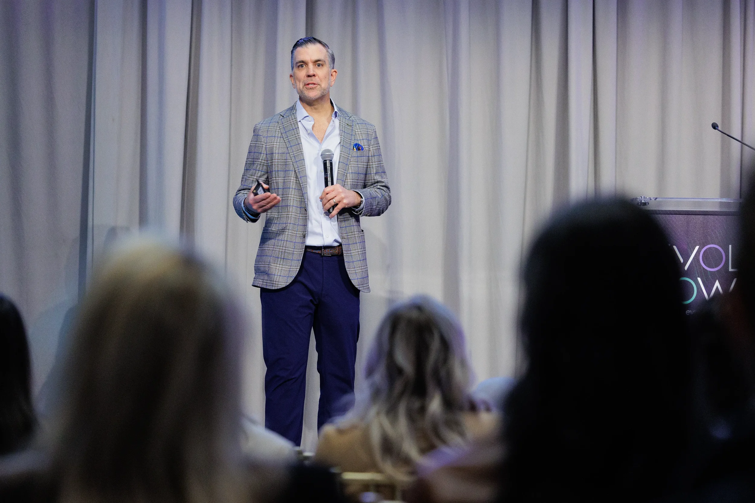 Speaker in plaid blazer presents with microphone as audience silhouettes appear in foreground at Chicago conference