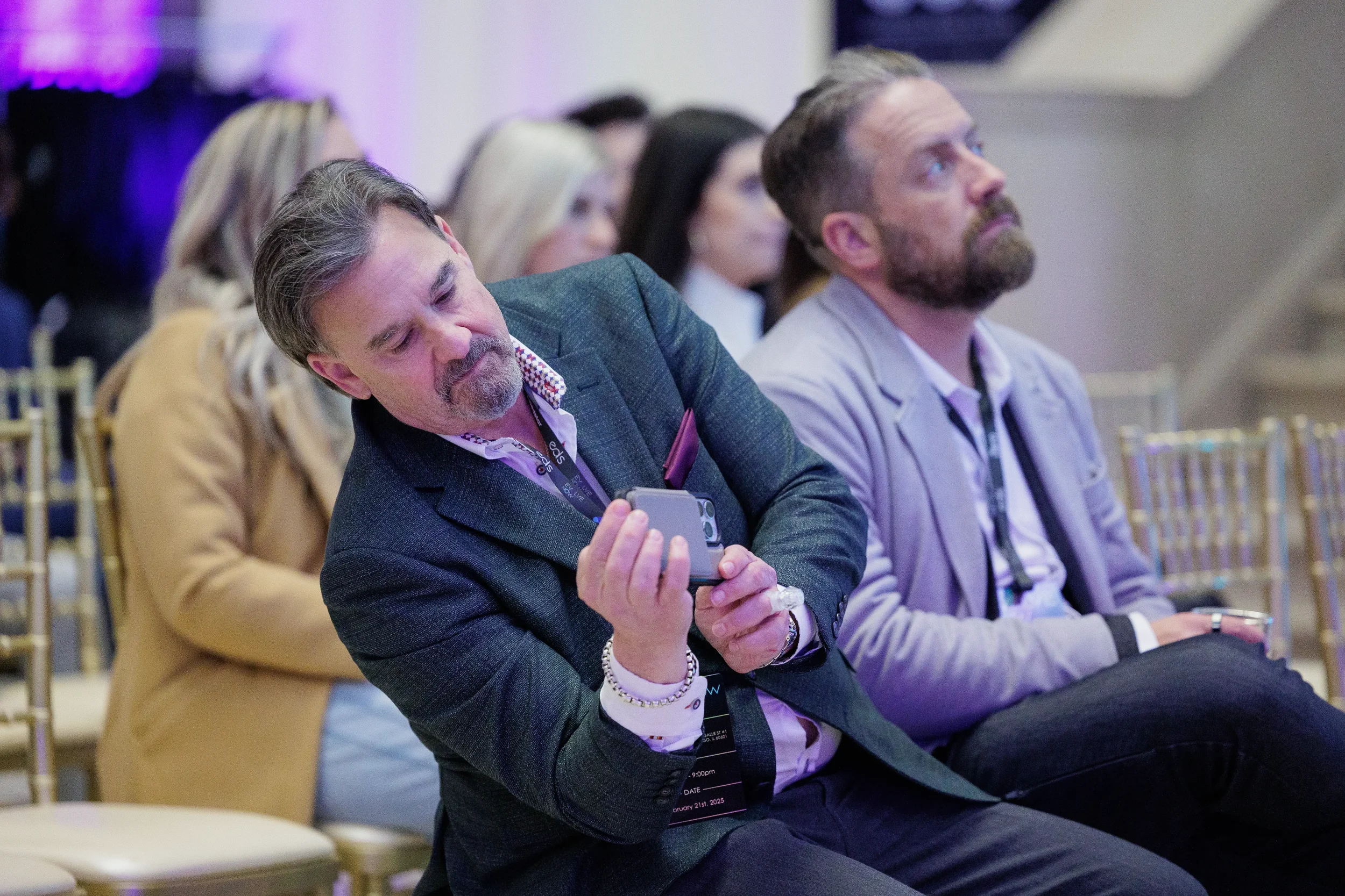 Two attendees sit in audience at Chicago conference, one checking phone during session