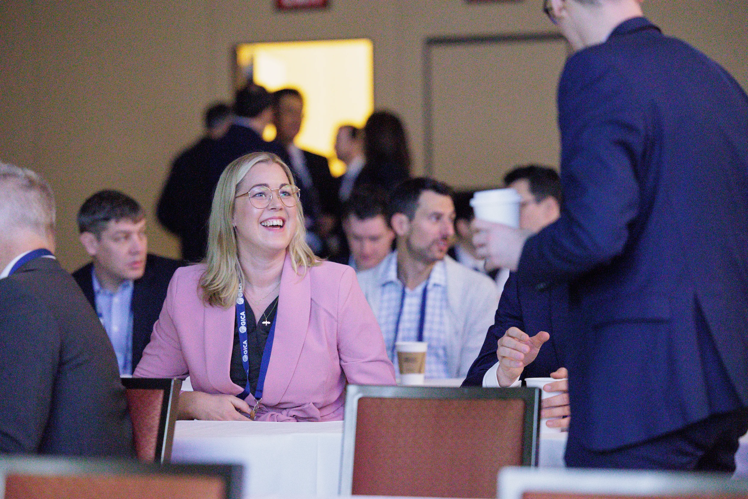 A smiling blonde woman in a pink blazer and glasses sits at a conference table in Orlando with other attendees.