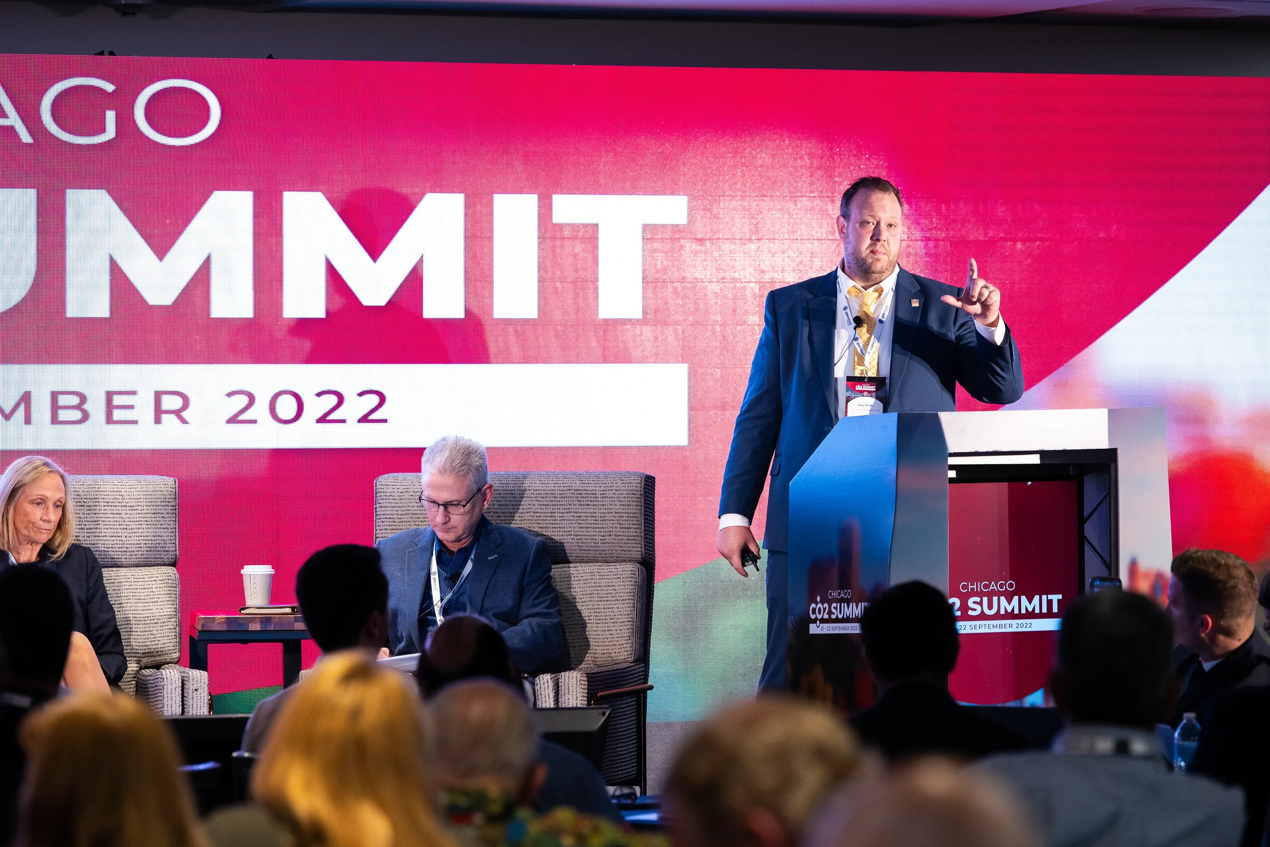 Speaker points toward audience from podium on stage in front of large branded backdrop at Chicago industry conference