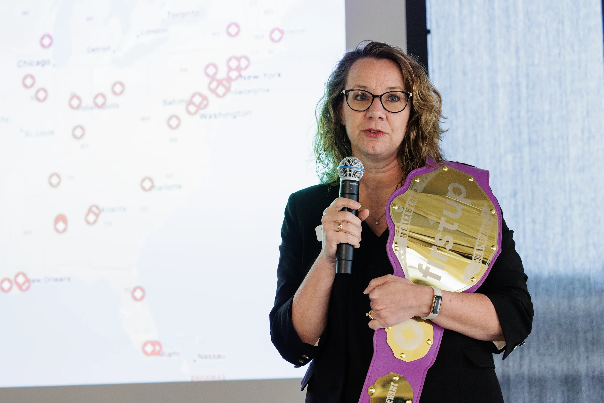 Female speaker holds a branded champion belt and microphone beside a map slide at Chicago conference