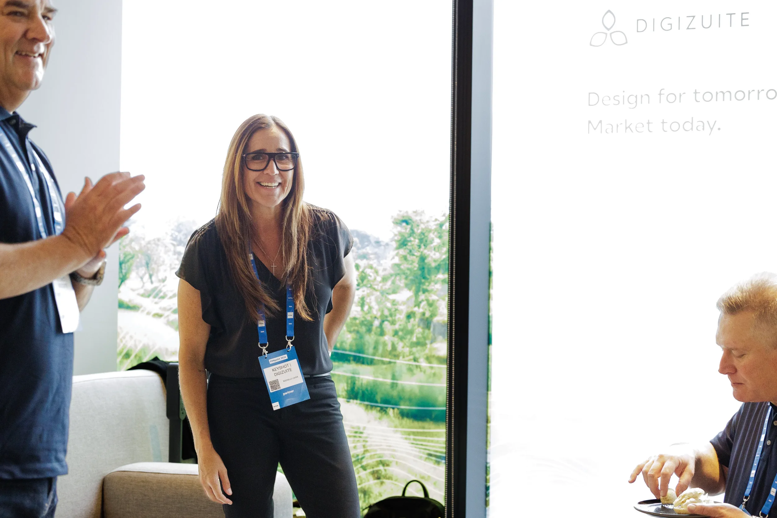 Smiling attendee with lanyard stands near window as colleague applauds at Orlando corporate event