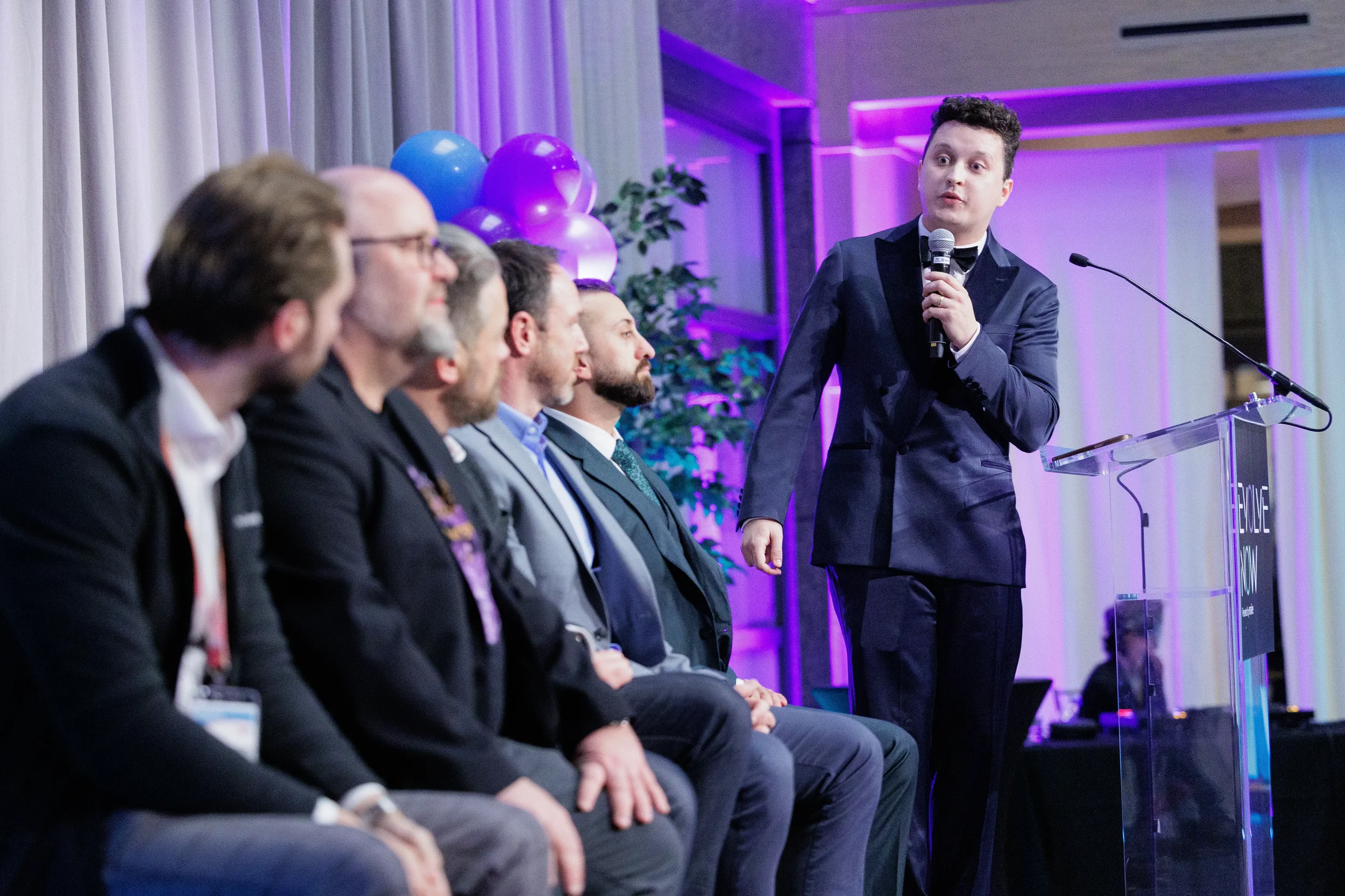 Tuxedo-clad emcee addresses seated panel at acrylic podium with balloon décor at Orlando industry conference