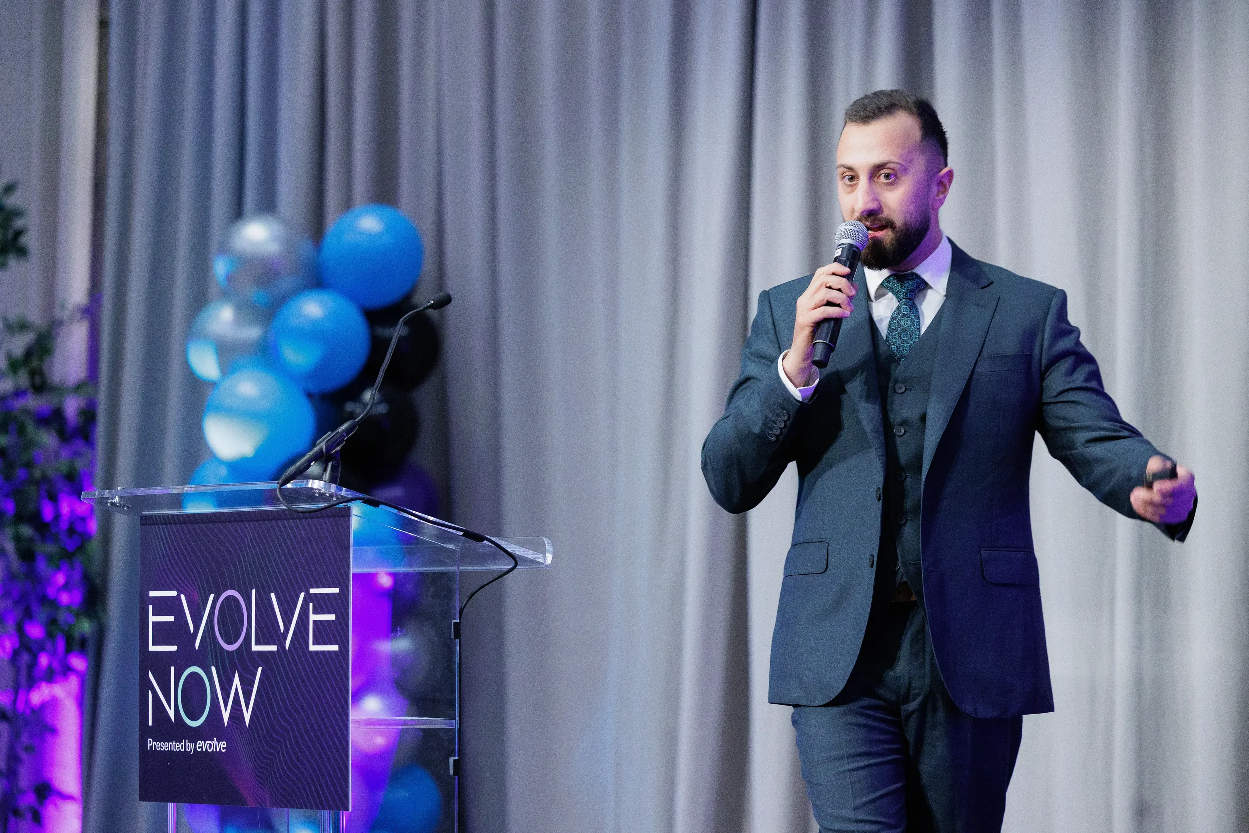 Three-piece suited speaker holds microphone beside branded podium with balloon column at Orlando corporate event