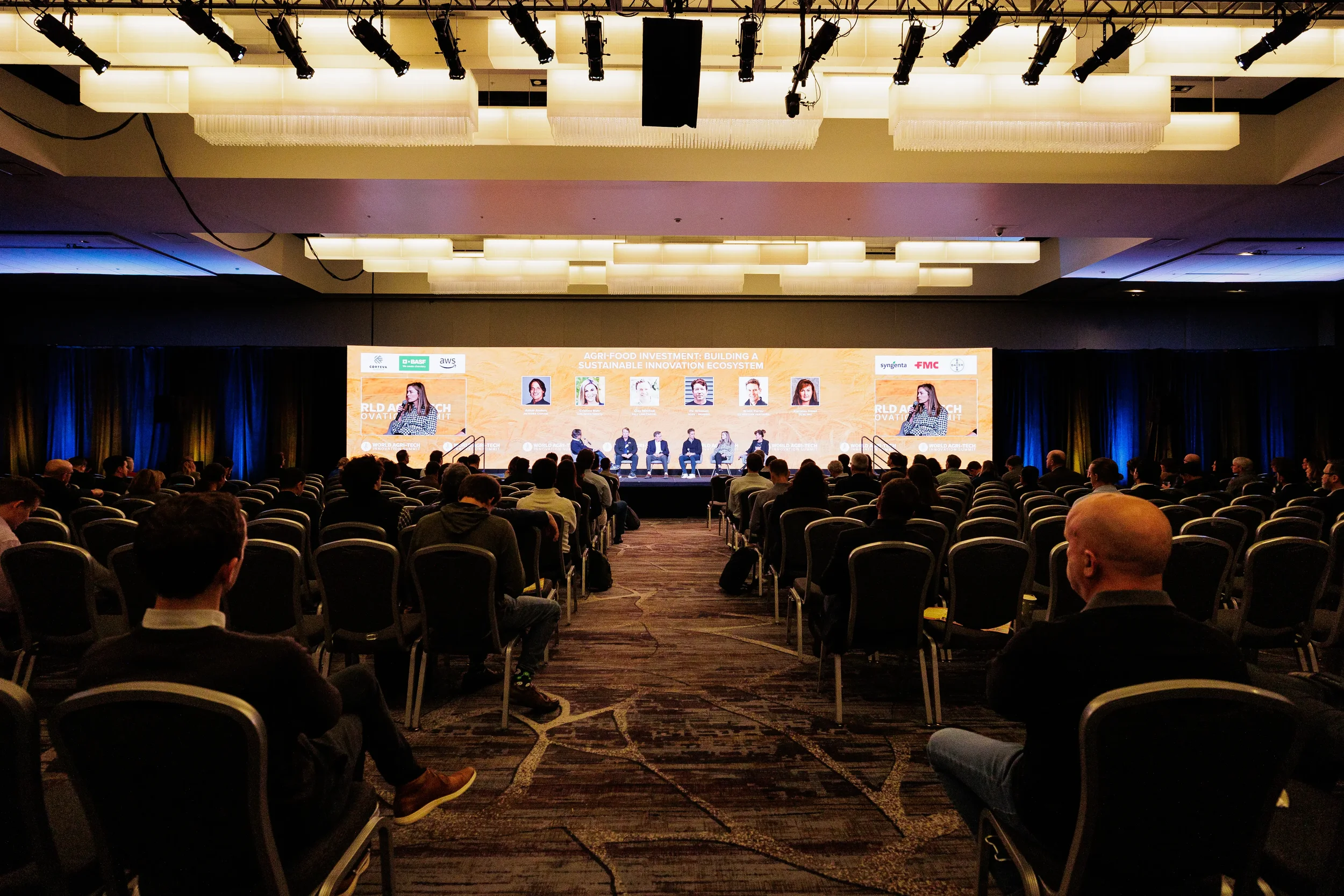 Wide shot of ballroom audience facing panel on branded stage with dramatic lighting at Orlando corporate conference