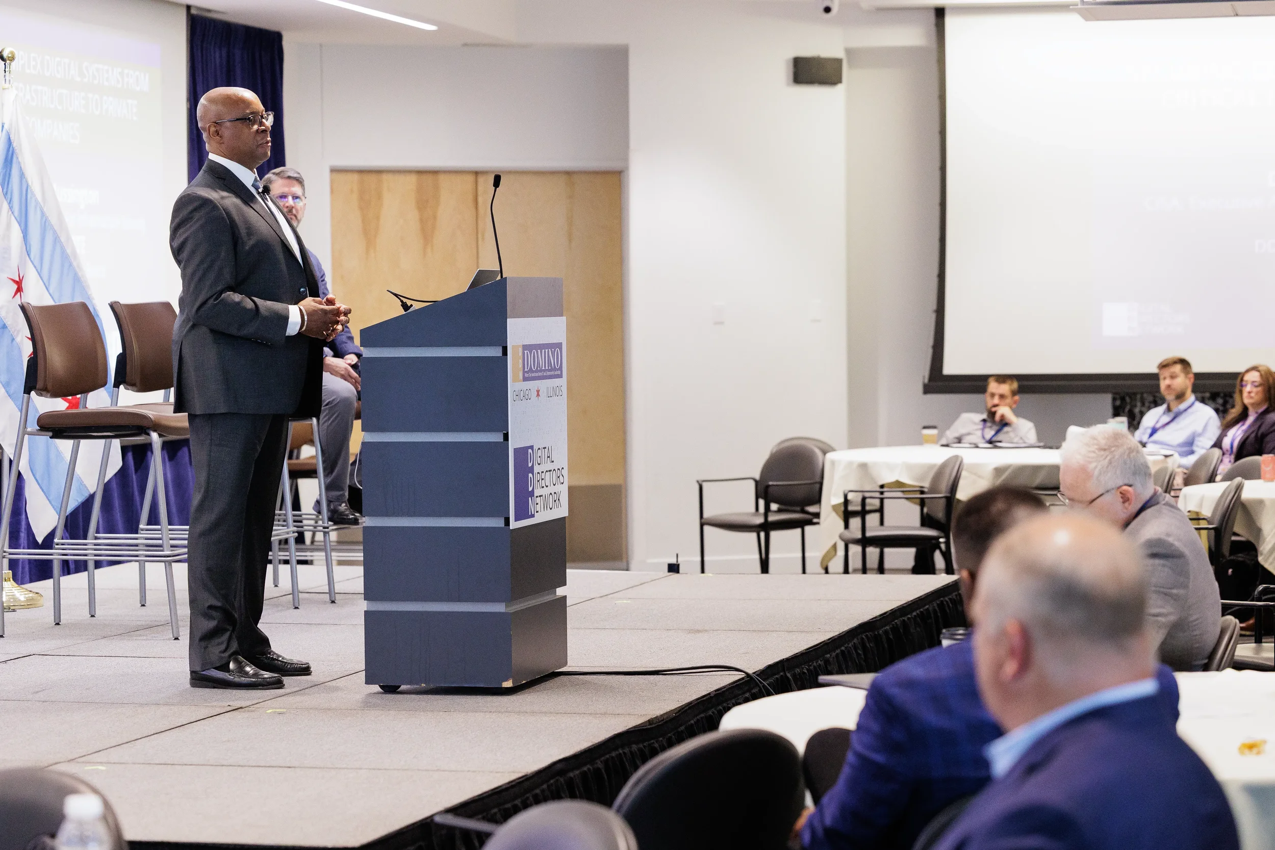 Speaker stands at branded podium on stage beside Chicago flag at Orlando corporate conference