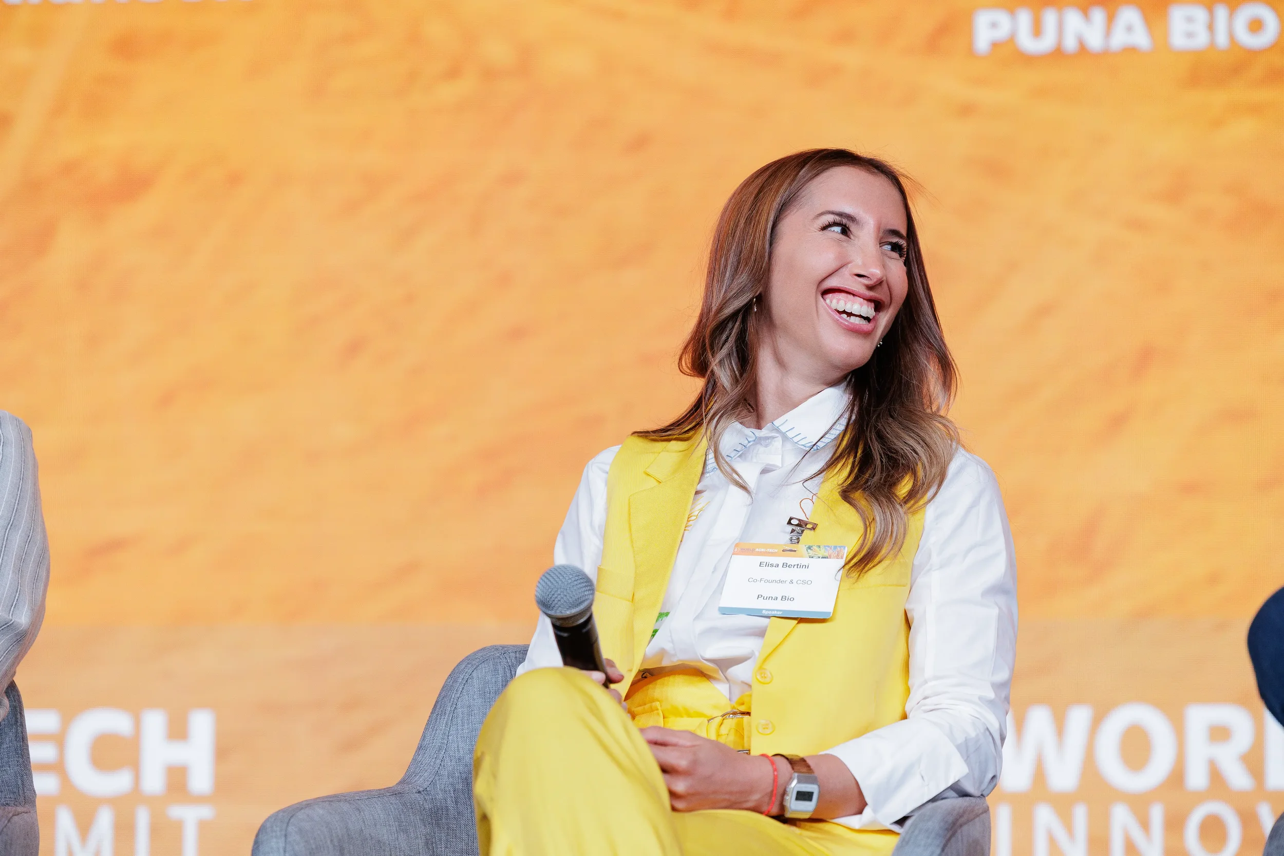 Panelist in yellow suit laughs while seated on stage with orange branded backdrop at Chicago conference