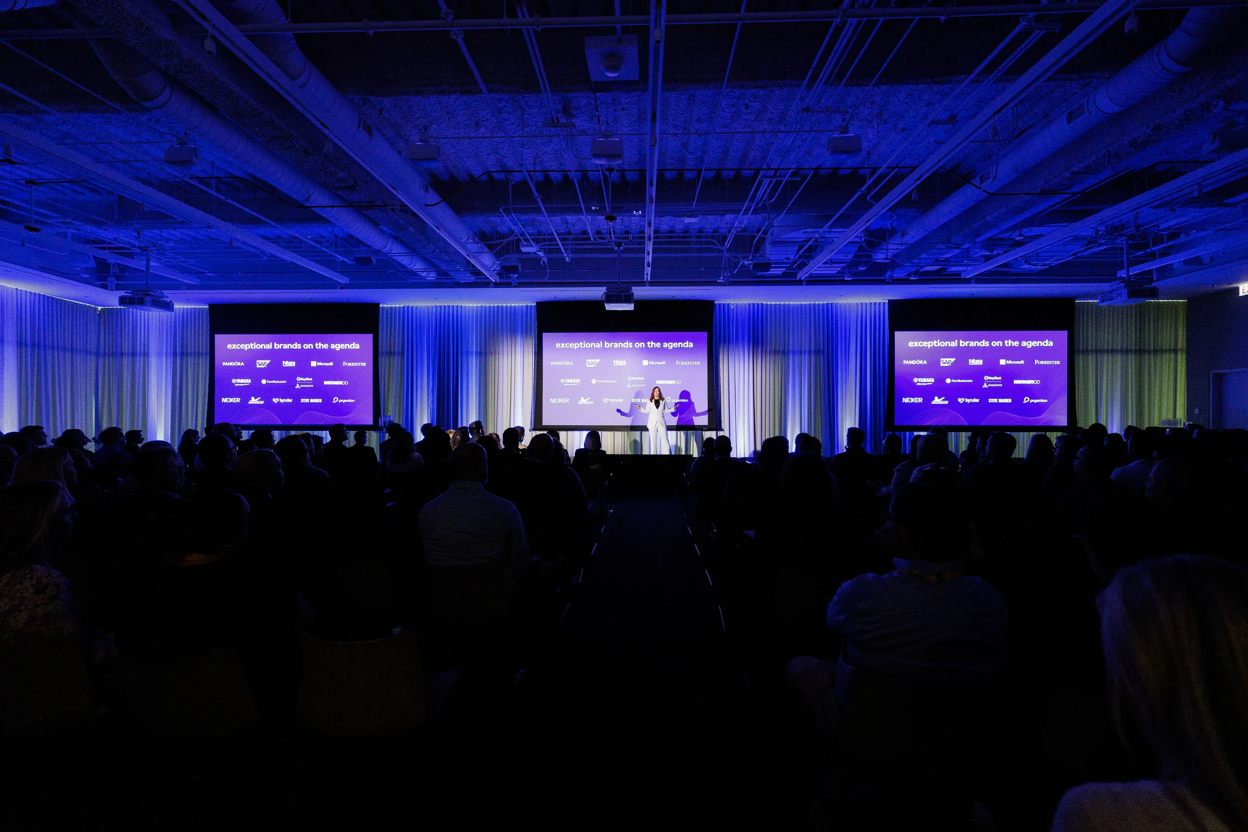 Speaker in white suit presents on stage before three screens and packed audience at Chicago corporate conference