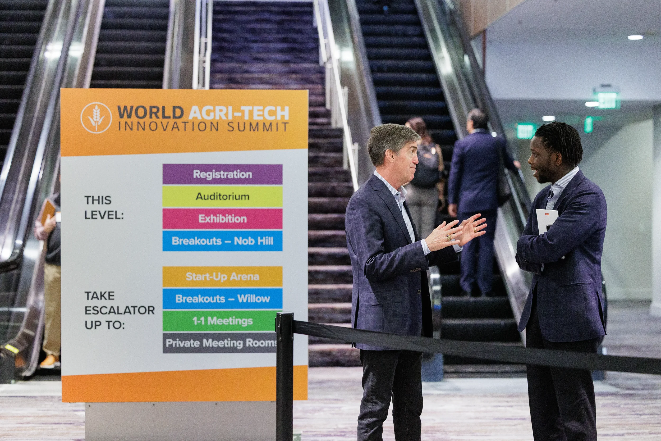 Two badged attendees converse beside branded wayfinding sign near escalator at Chicago corporate conference venue