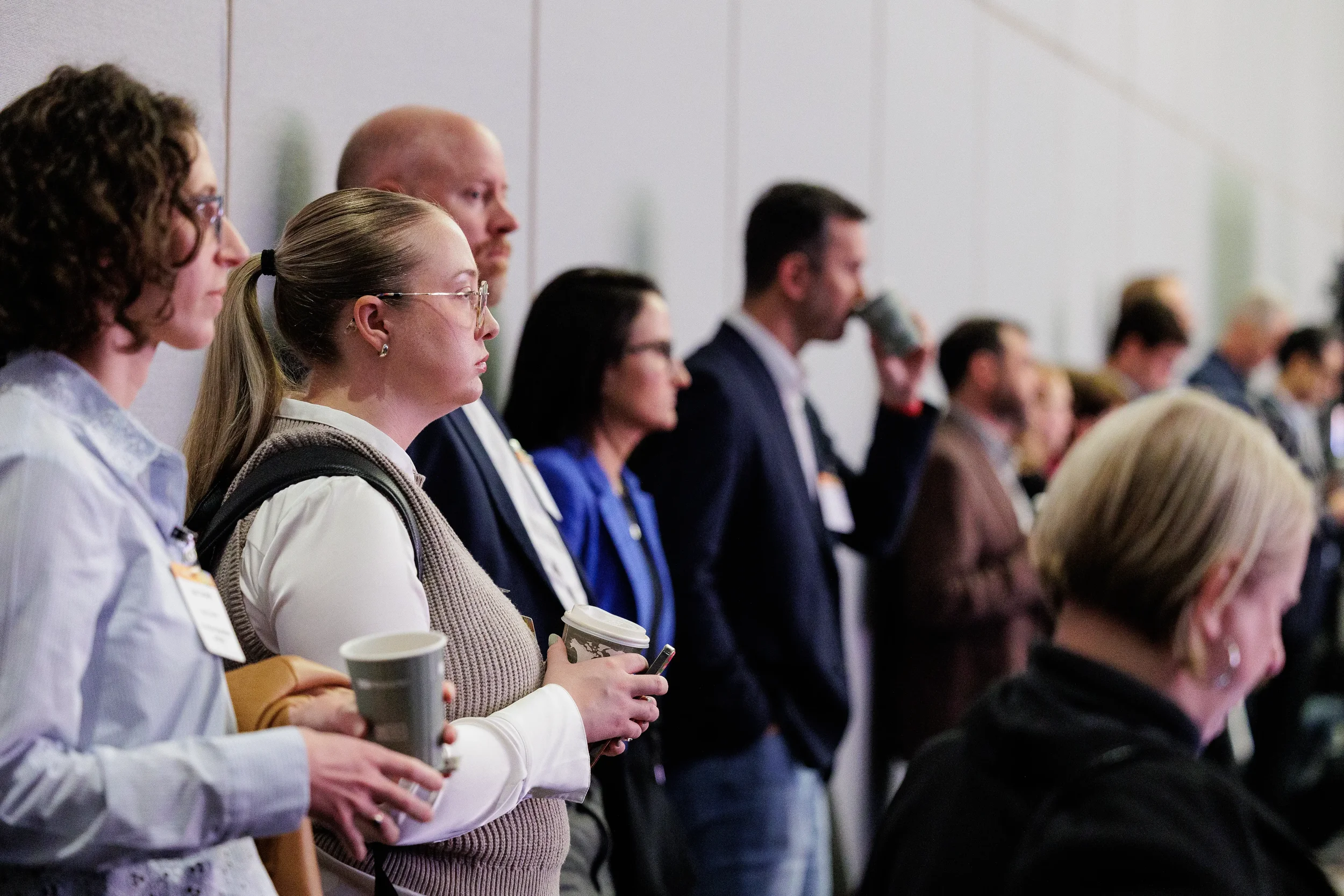 Row of badged attendees with coffee cups standing along wall during Orlando corporate event session