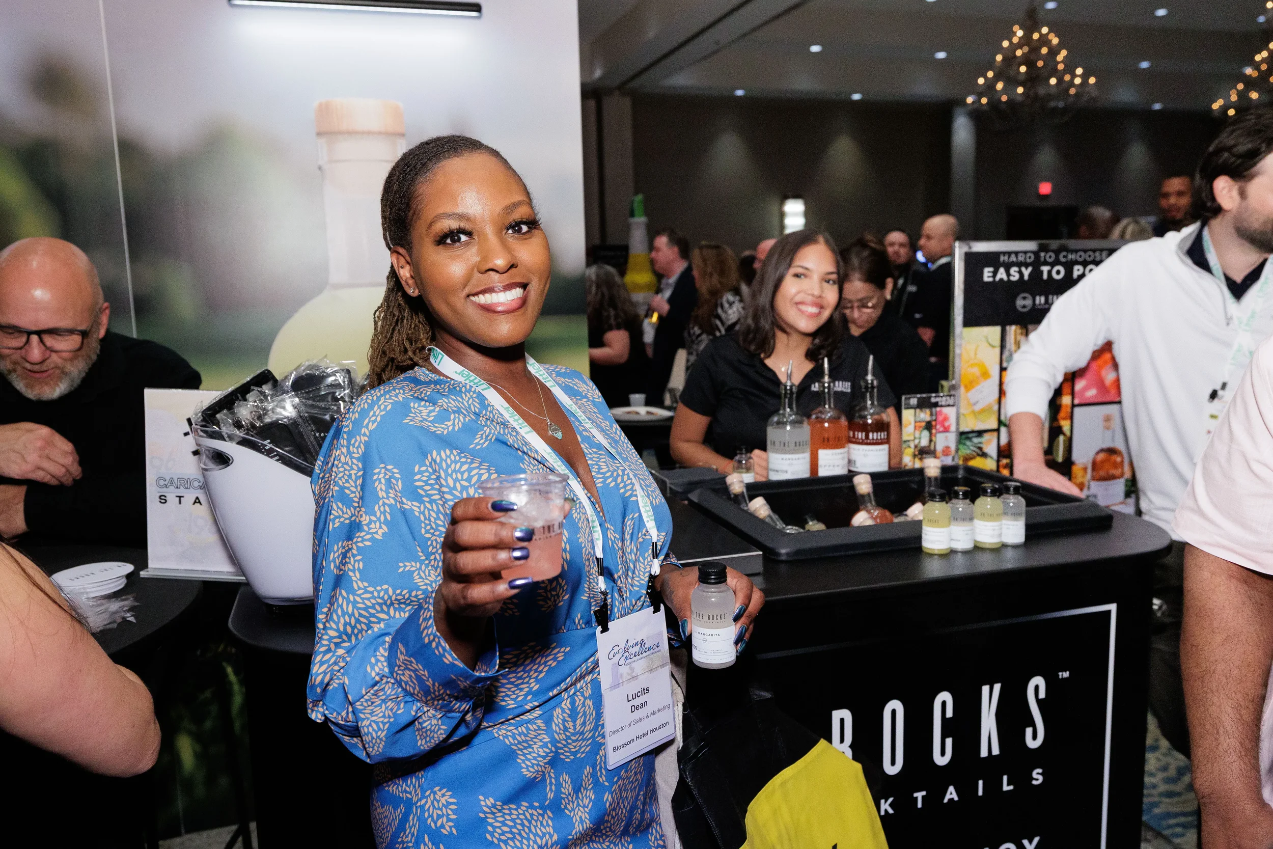 Smiling female attendee with lanyard holds drink at sponsor cocktail booth during Orlando corporate event