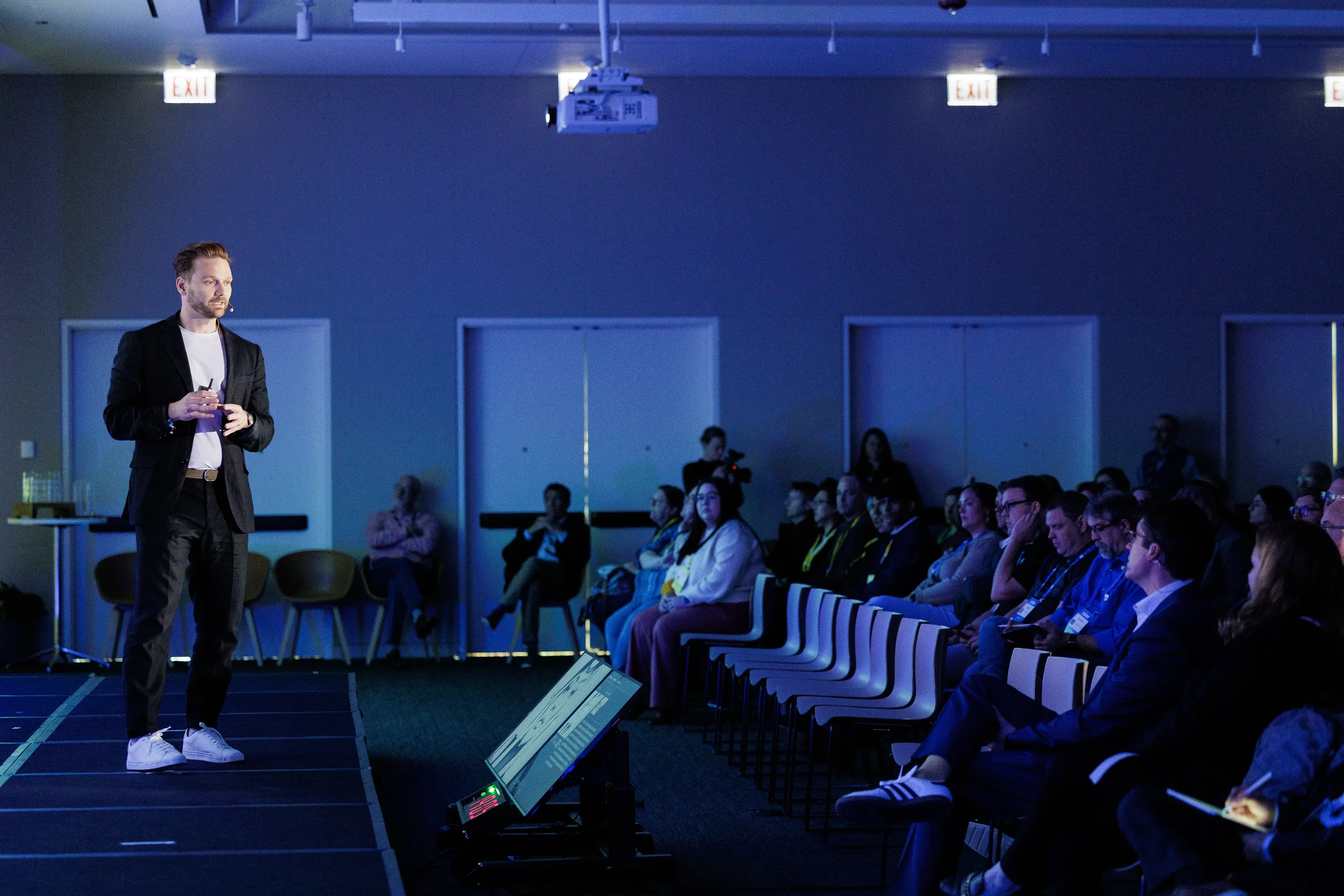 Speaker stands on stage facing seated audience in blue-lit room at Orlando industry conference