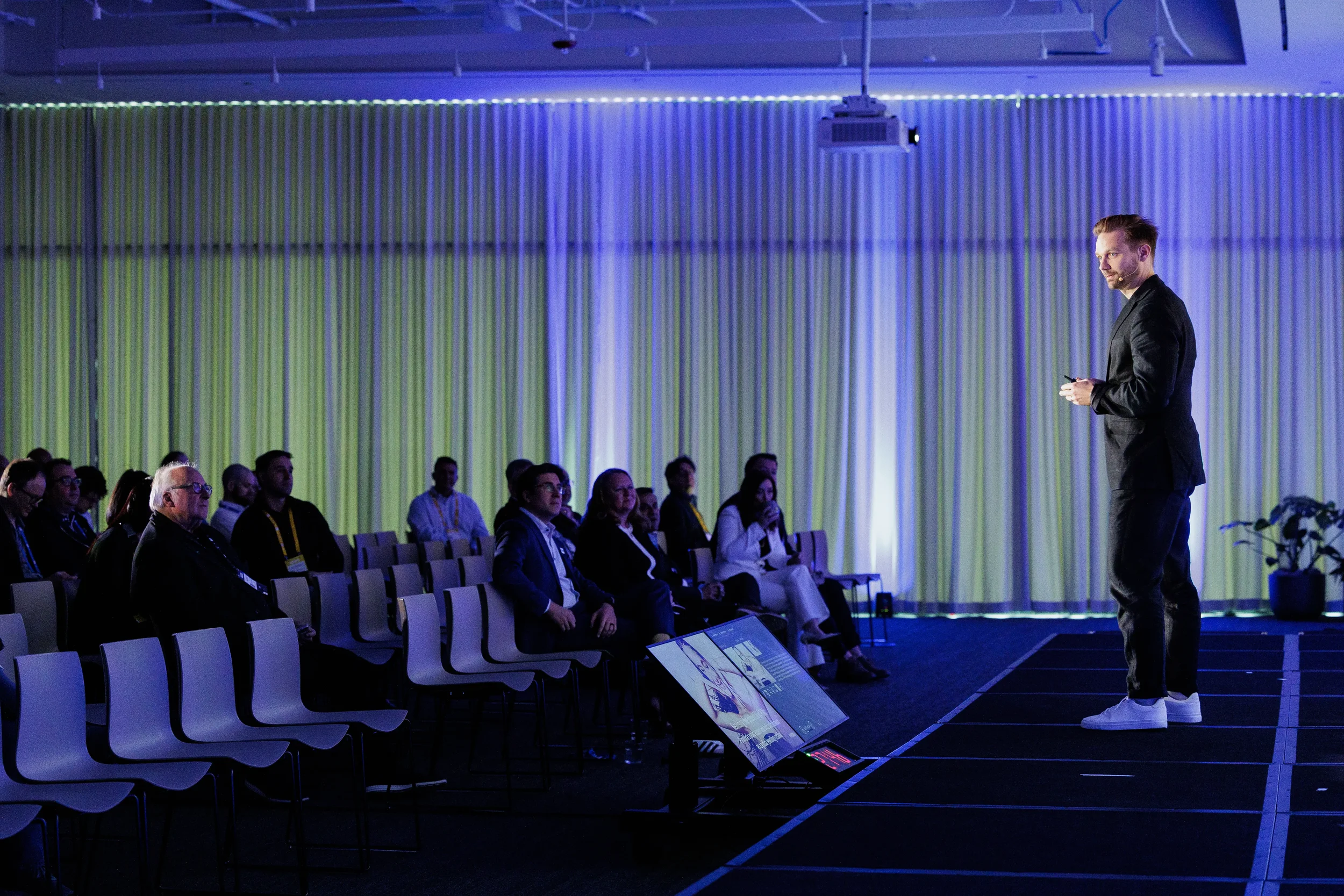 Speaker in dark suit addresses seated audience on stage in blue-lit room at Orlando conference