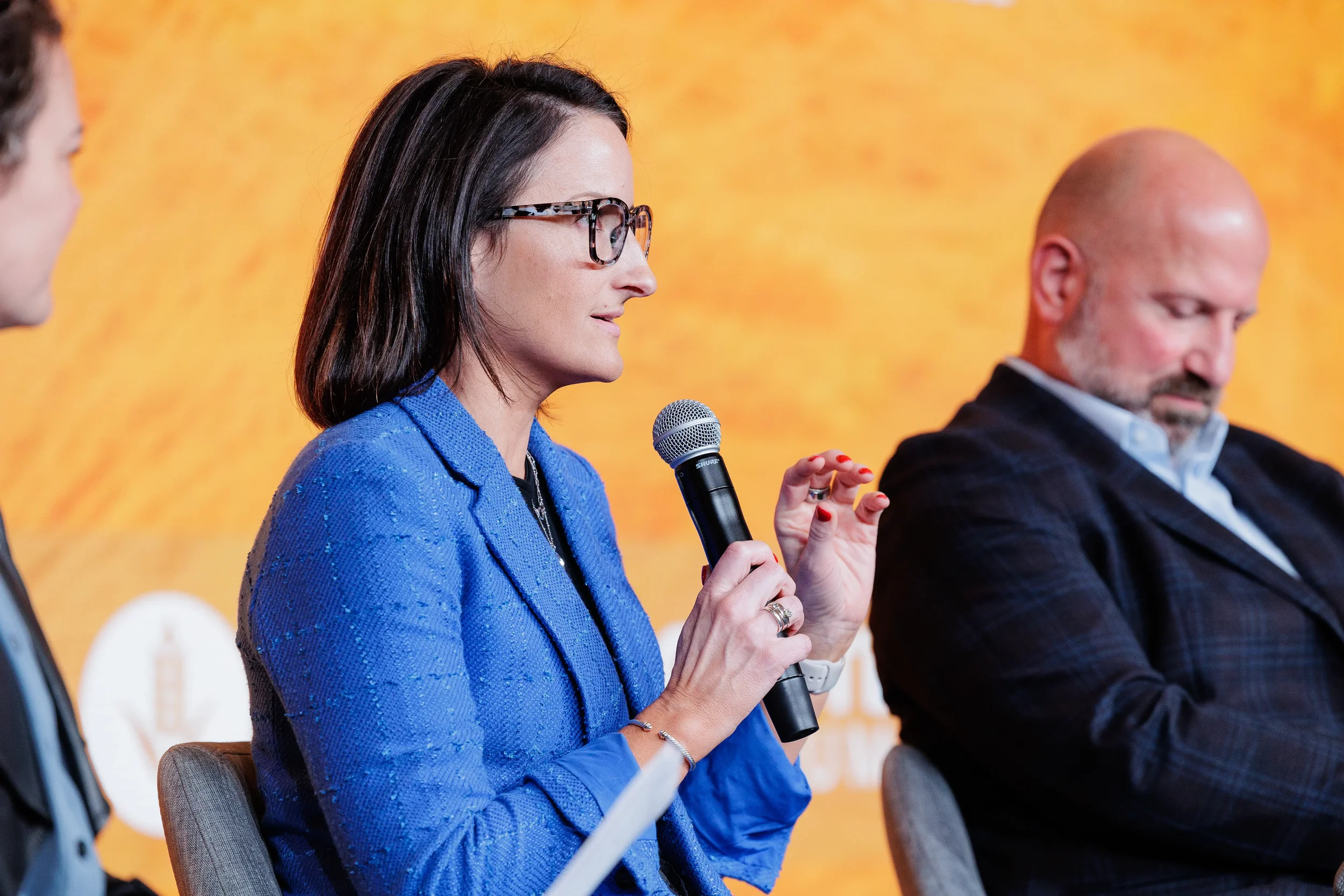 Female panelist in blue blazer holds microphone on stage before orange backdrop at Orlando industry conference
