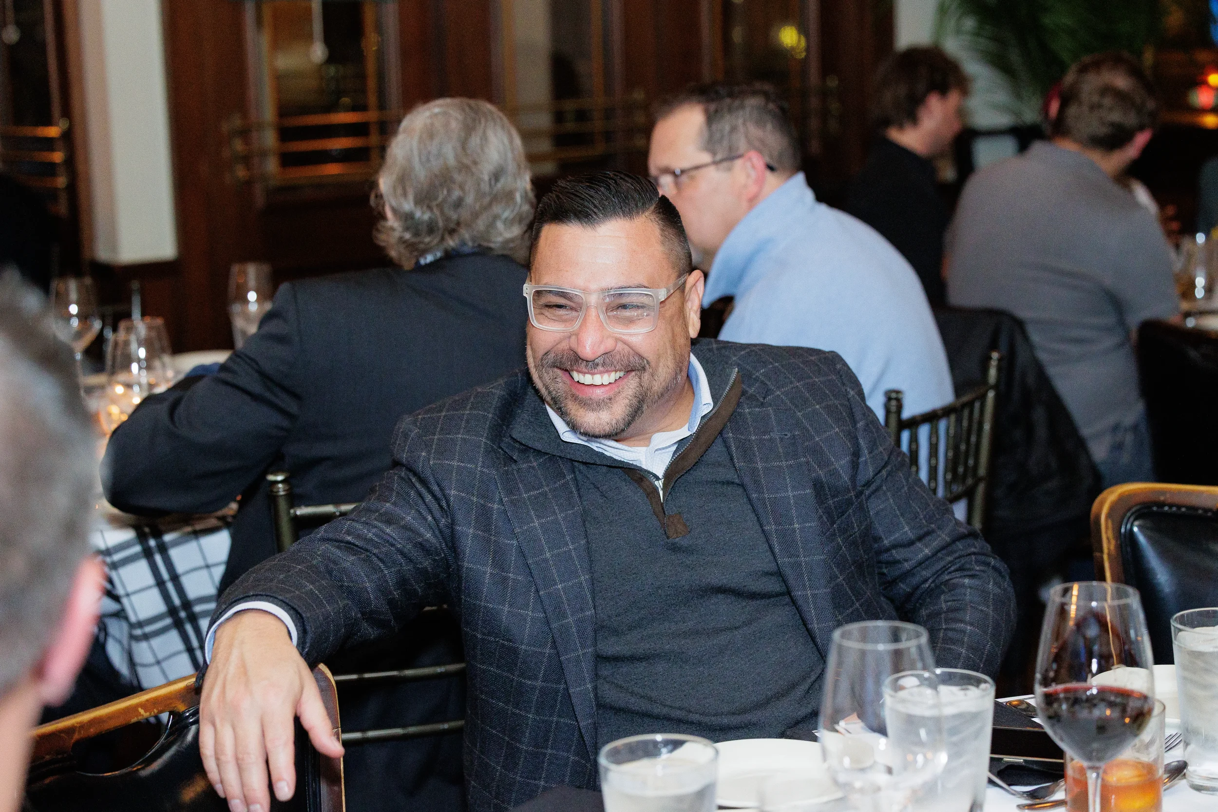 Smiling man in a plaid blazer and glasses sits at a dinner table after a conference in Orlando. Other guests sit nearby.