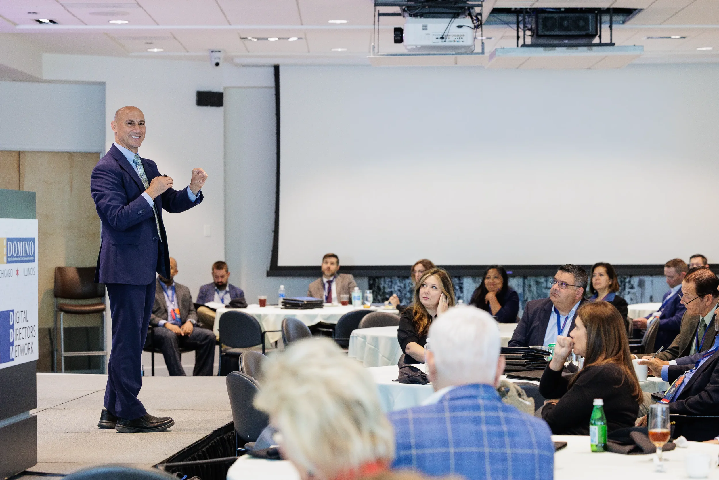 Male speaker gestures energetically on stage beside branded podium before engaged audience at Orlando corporate event