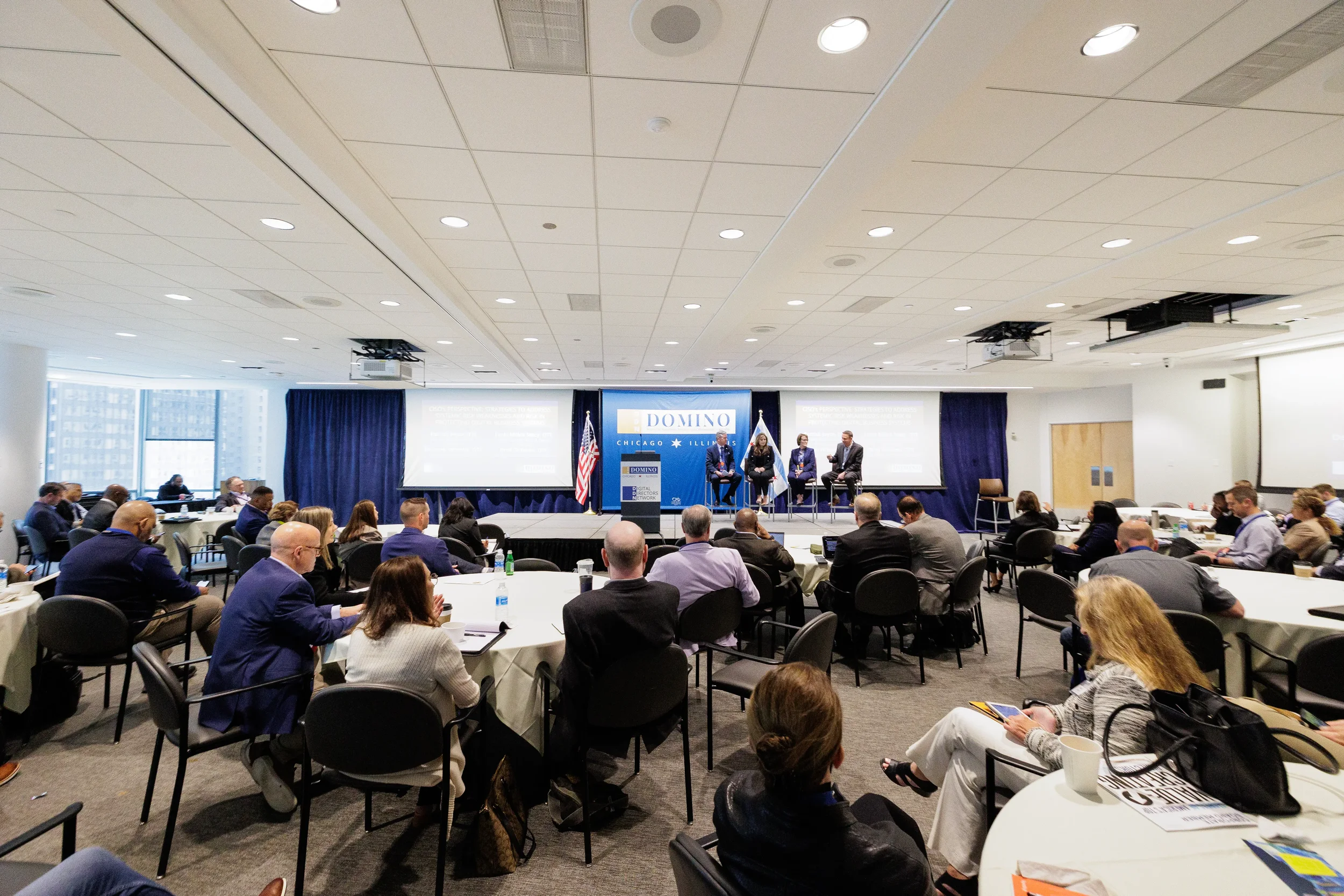 Three panelists on branded stage before full room of attendees at round tables at Orlando corporate event