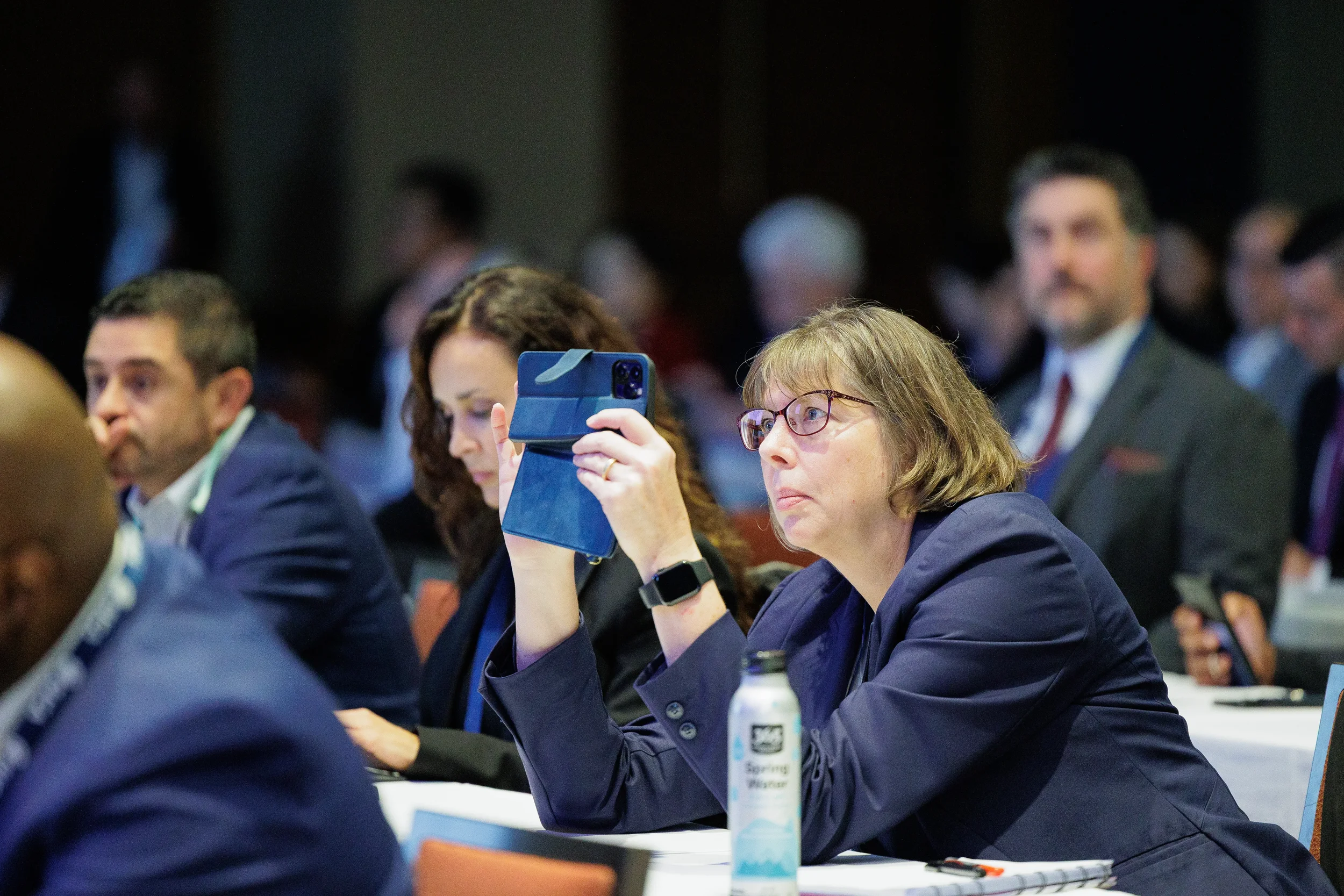 Woman in glasses wearing a navy blazer holds up a smartphone at a formal conference in Orlando.