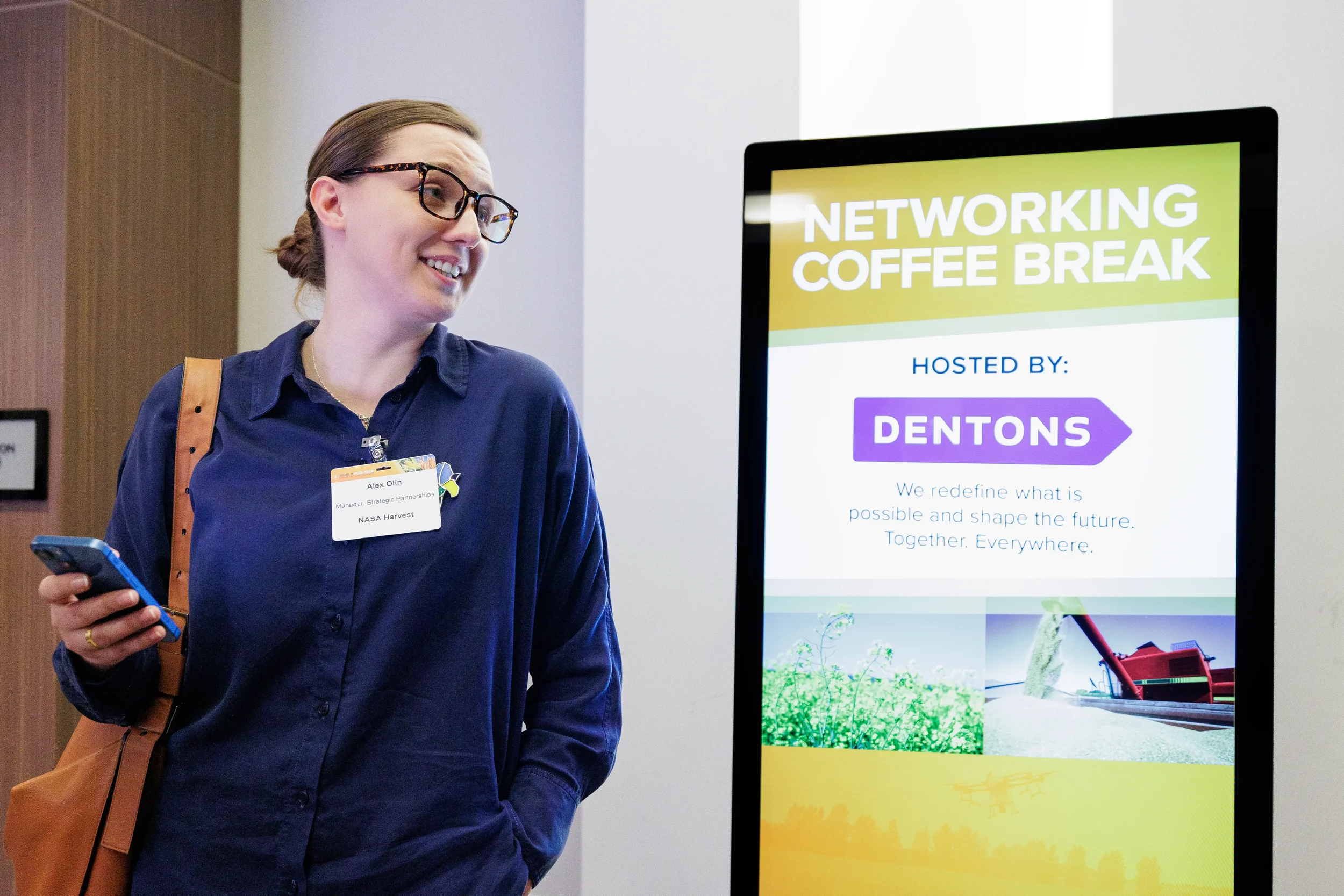 Smiling badged attendee with phone stands beside digital networking break signage at Orlando corporate event
