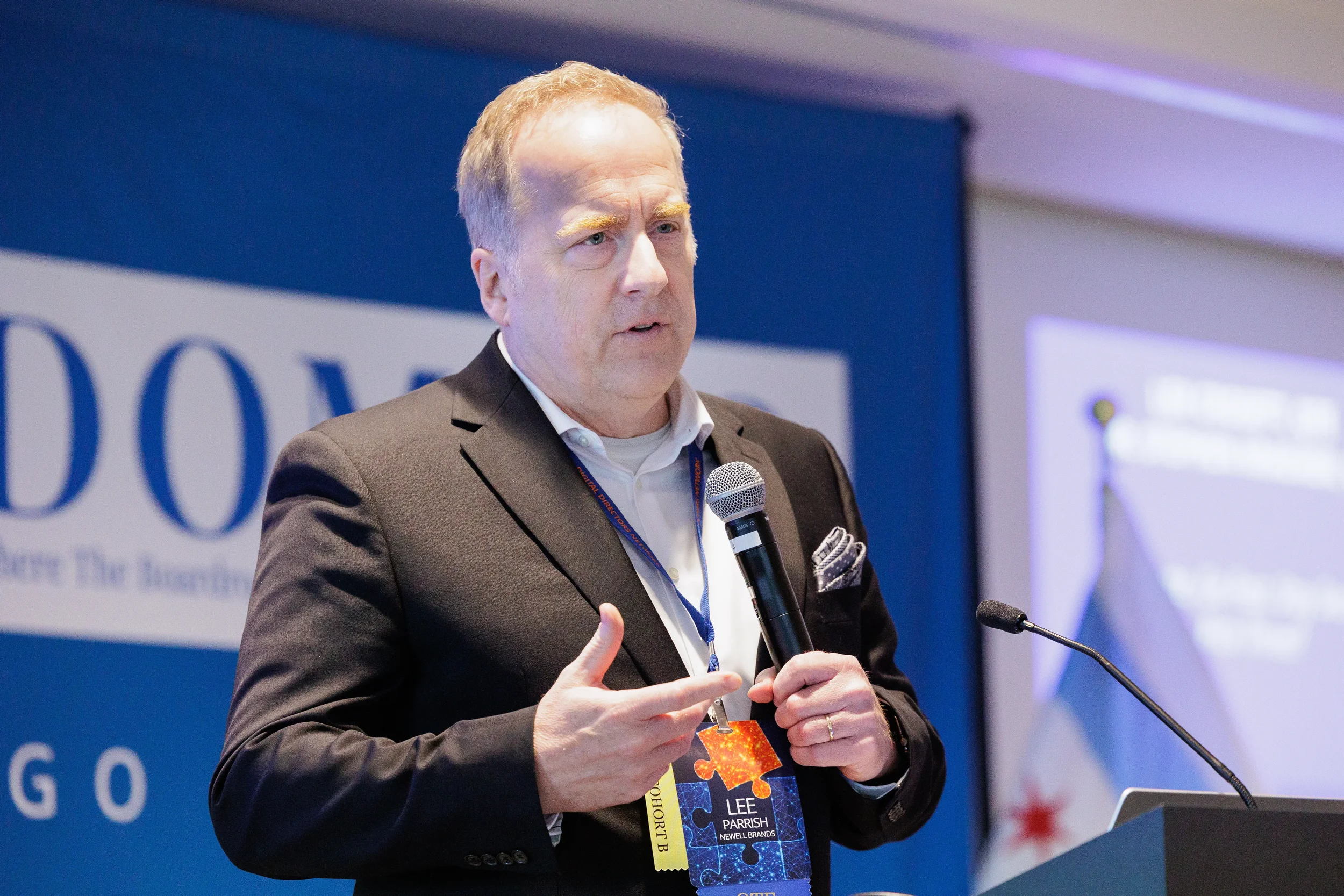 Male speaker holds microphone and gestures in front of branded backdrop at Orlando industry conference