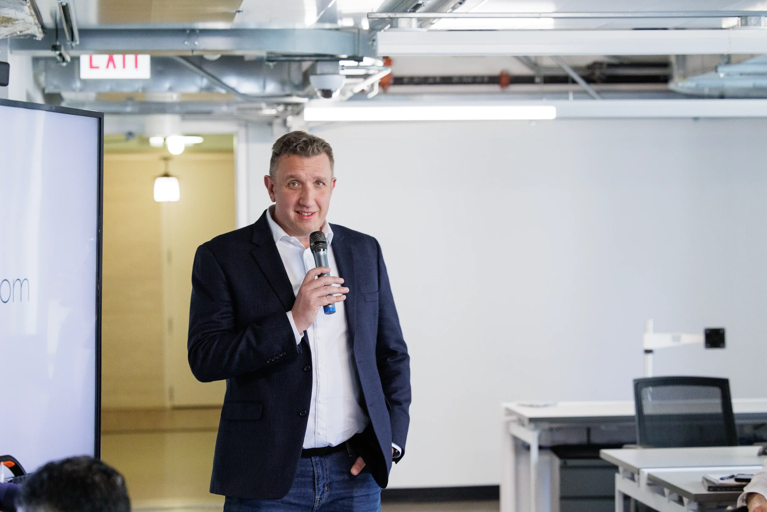 Male speaker presents with microphone beside a display screen in a modern space at Orlando conference