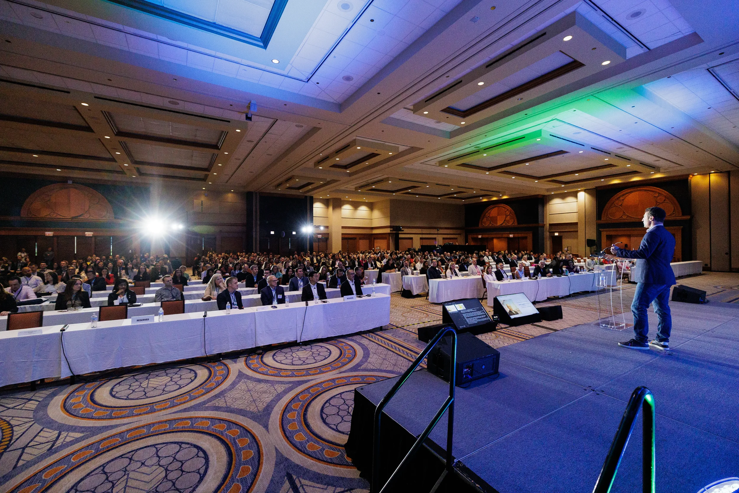 Packed ballroom audience at long tables listens to speaker on stage at Orlando corporate meeting