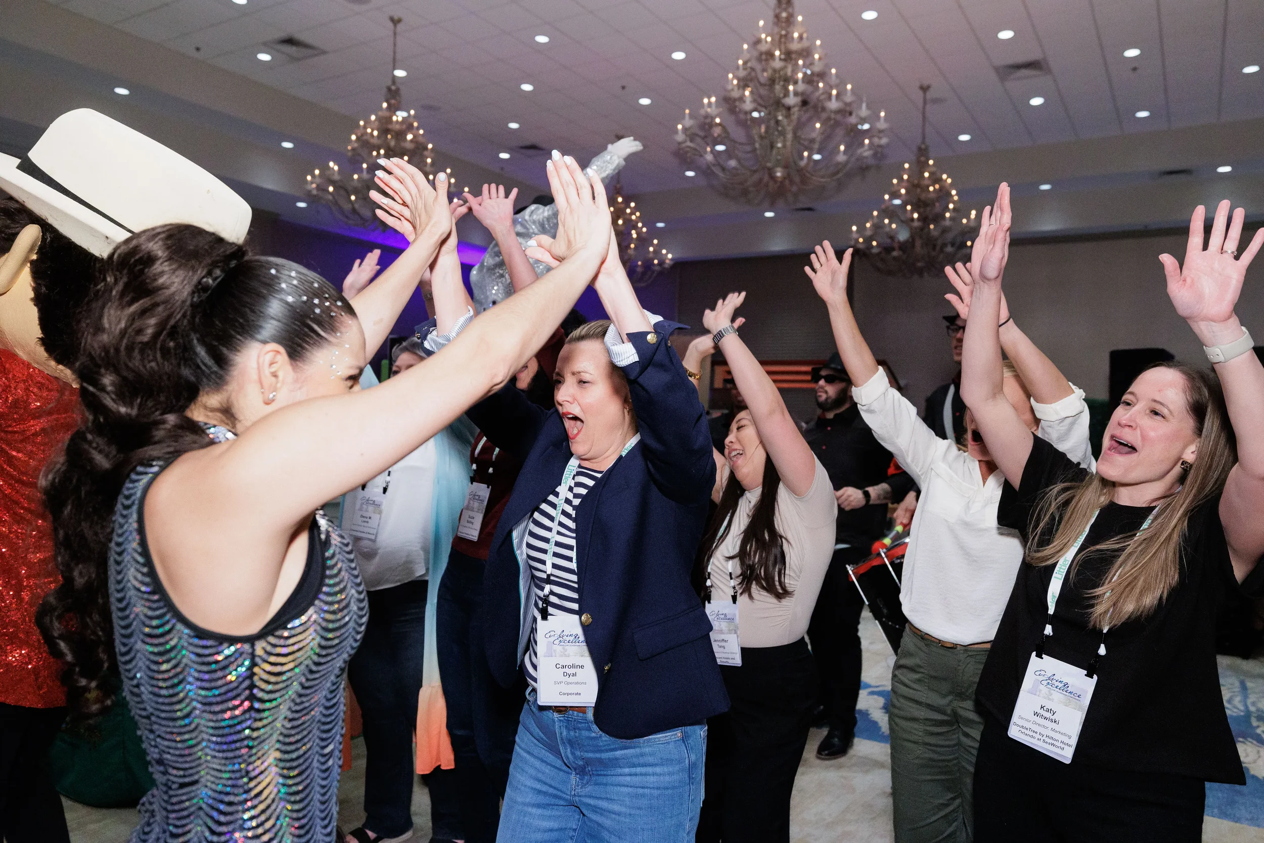 Attendees with lanyards dance with hands raised alongside performer in chandelier ballroom at Orlando conference