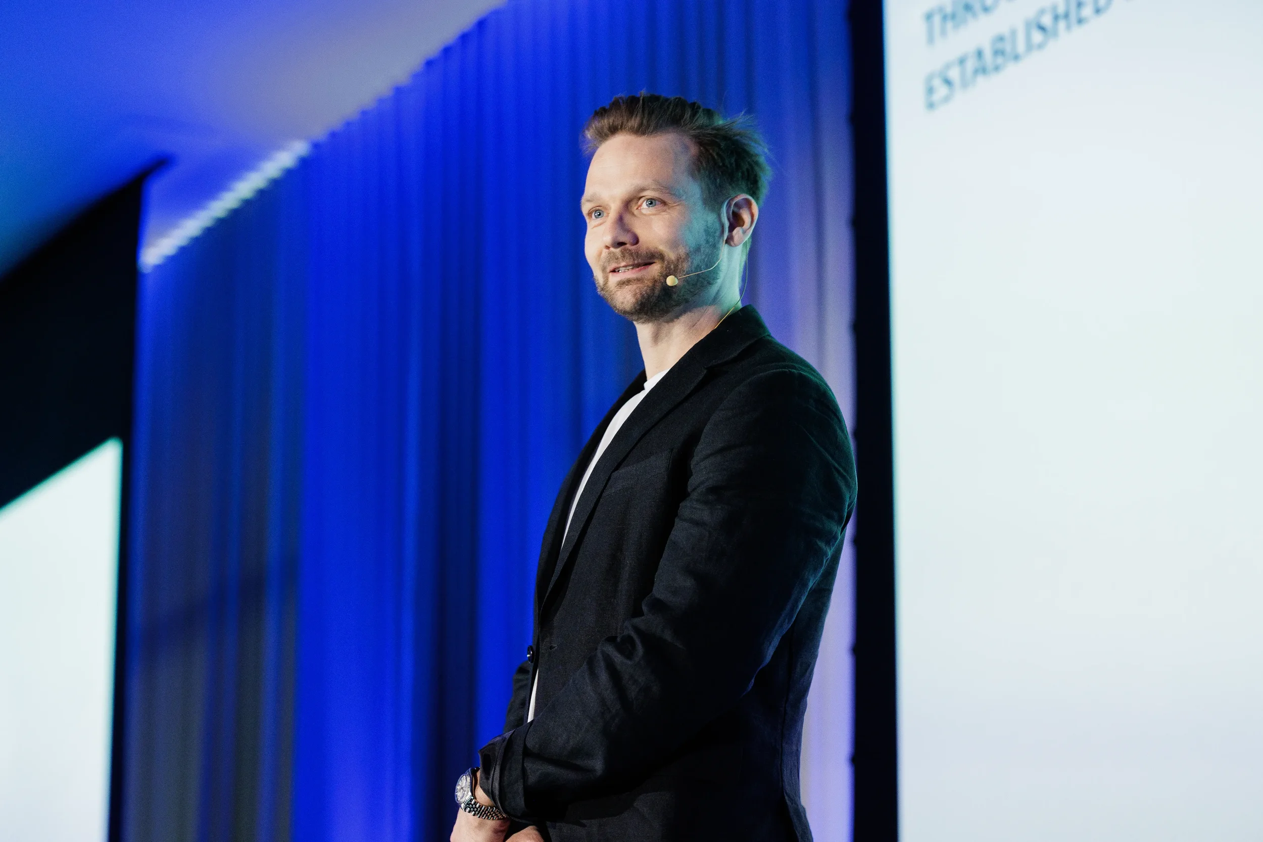 Speaker in black blazer with headset microphone smiles on stage by blue curtain at Orlando conference