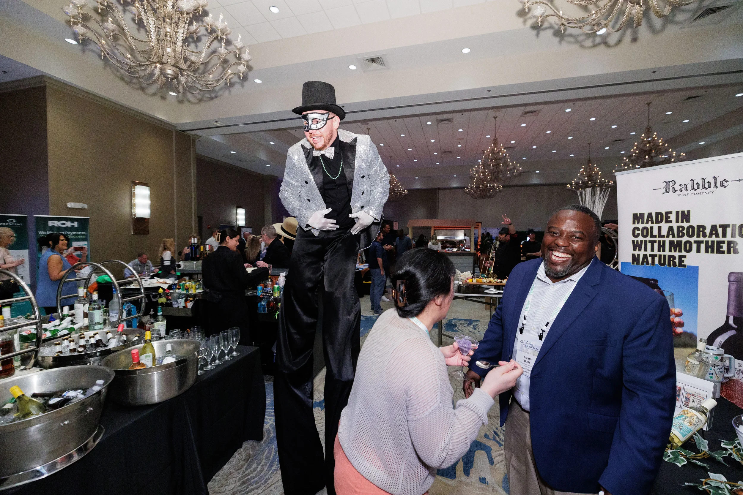Stilt performer in top hat entertains laughing attendees near wine booth at Orlando corporate conference expo