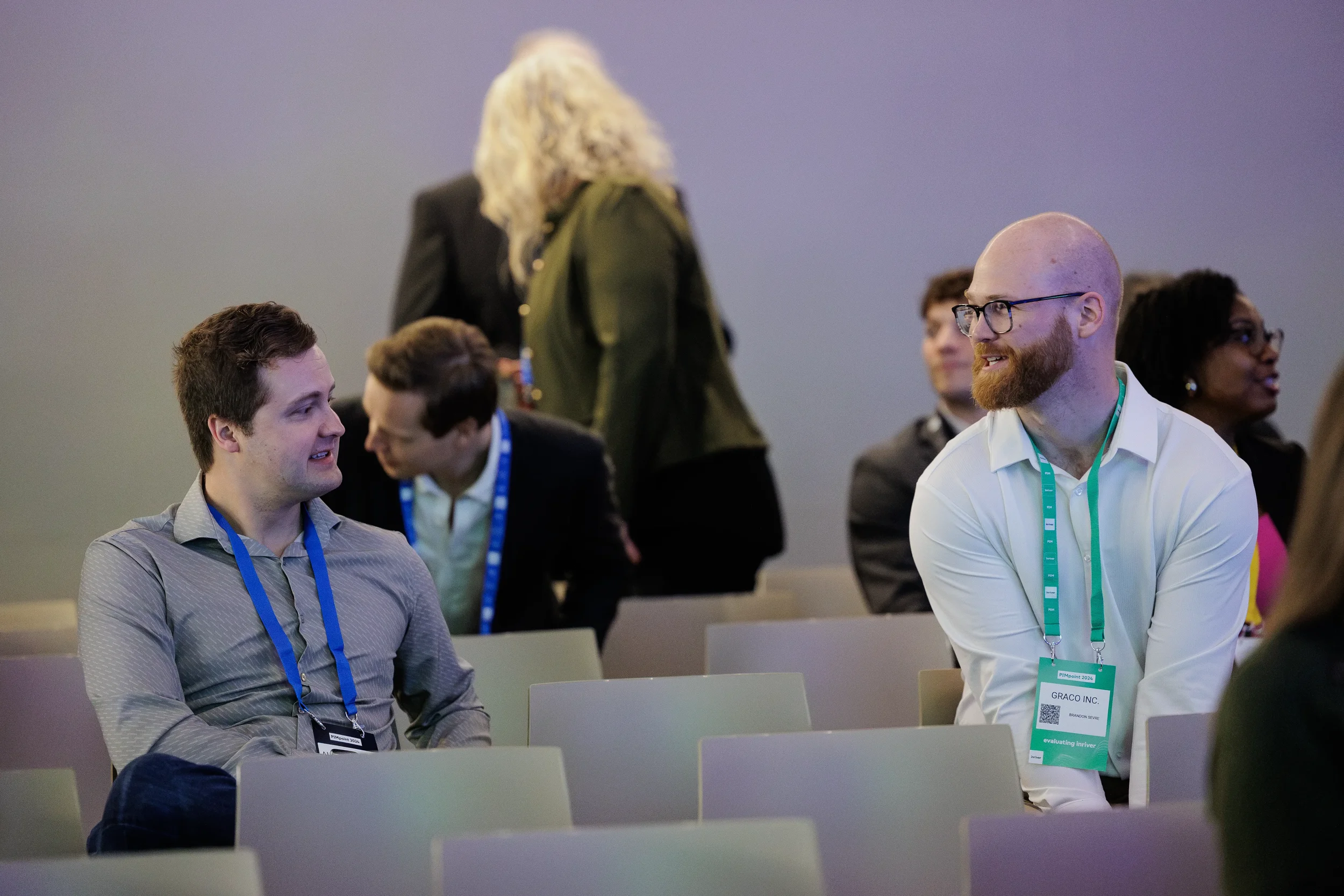 Two attendees with lanyards chat in seated audience before session at Orlando industry conference