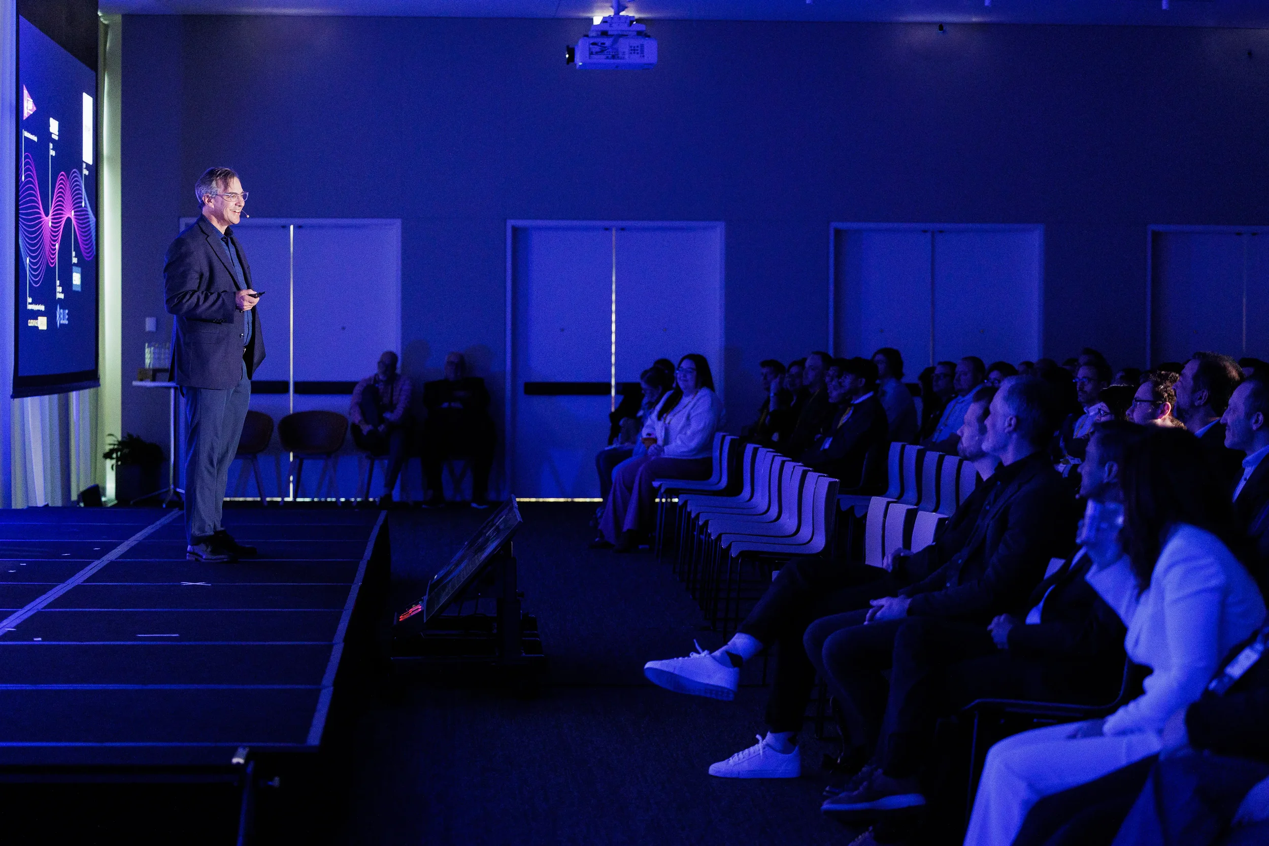 Smiling speaker on stage faces seated audience in blue-lit room at Chicago industry conference