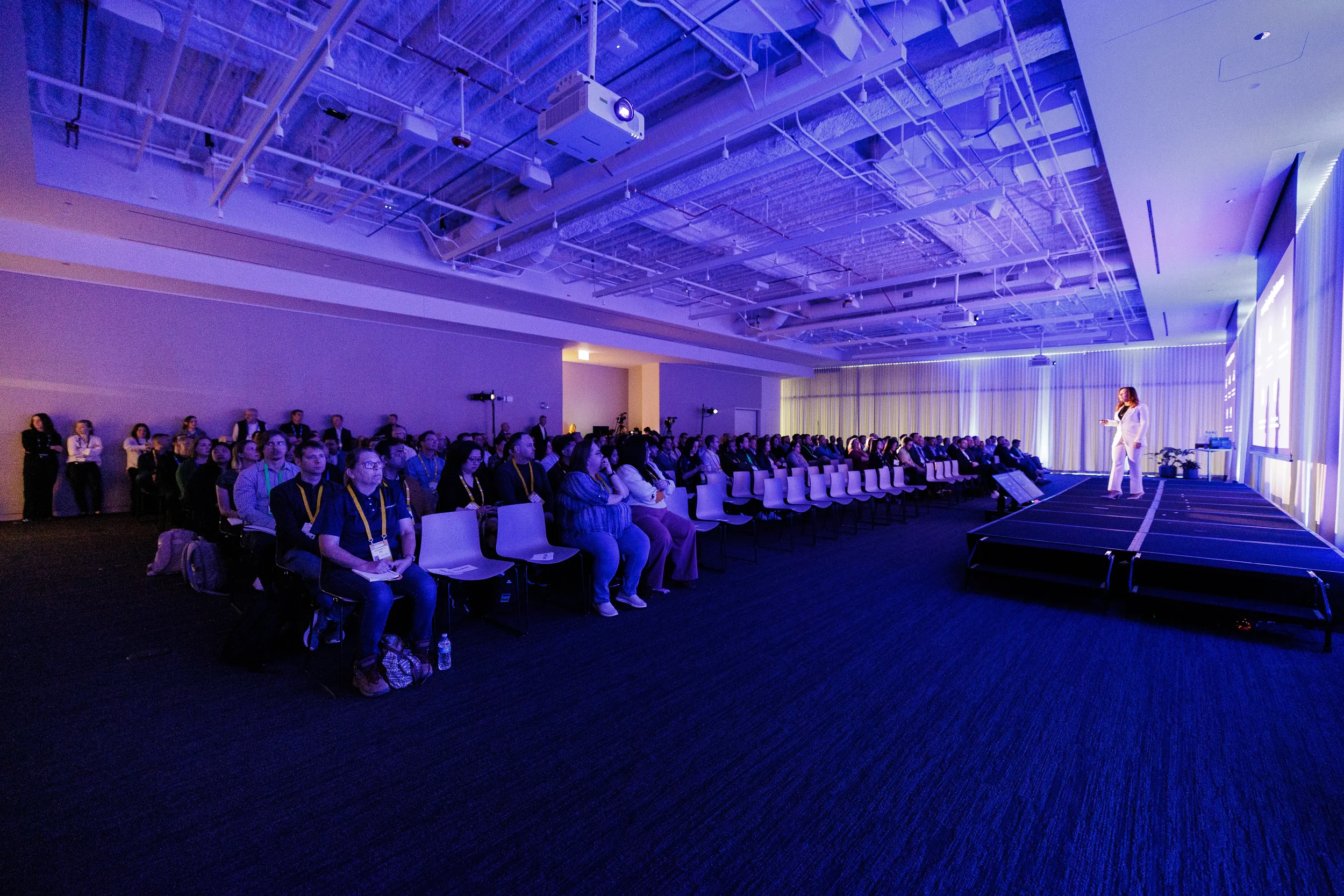 Speaker in white suit presents to seated audience in purple-lit ballroom at Orlando industry conference