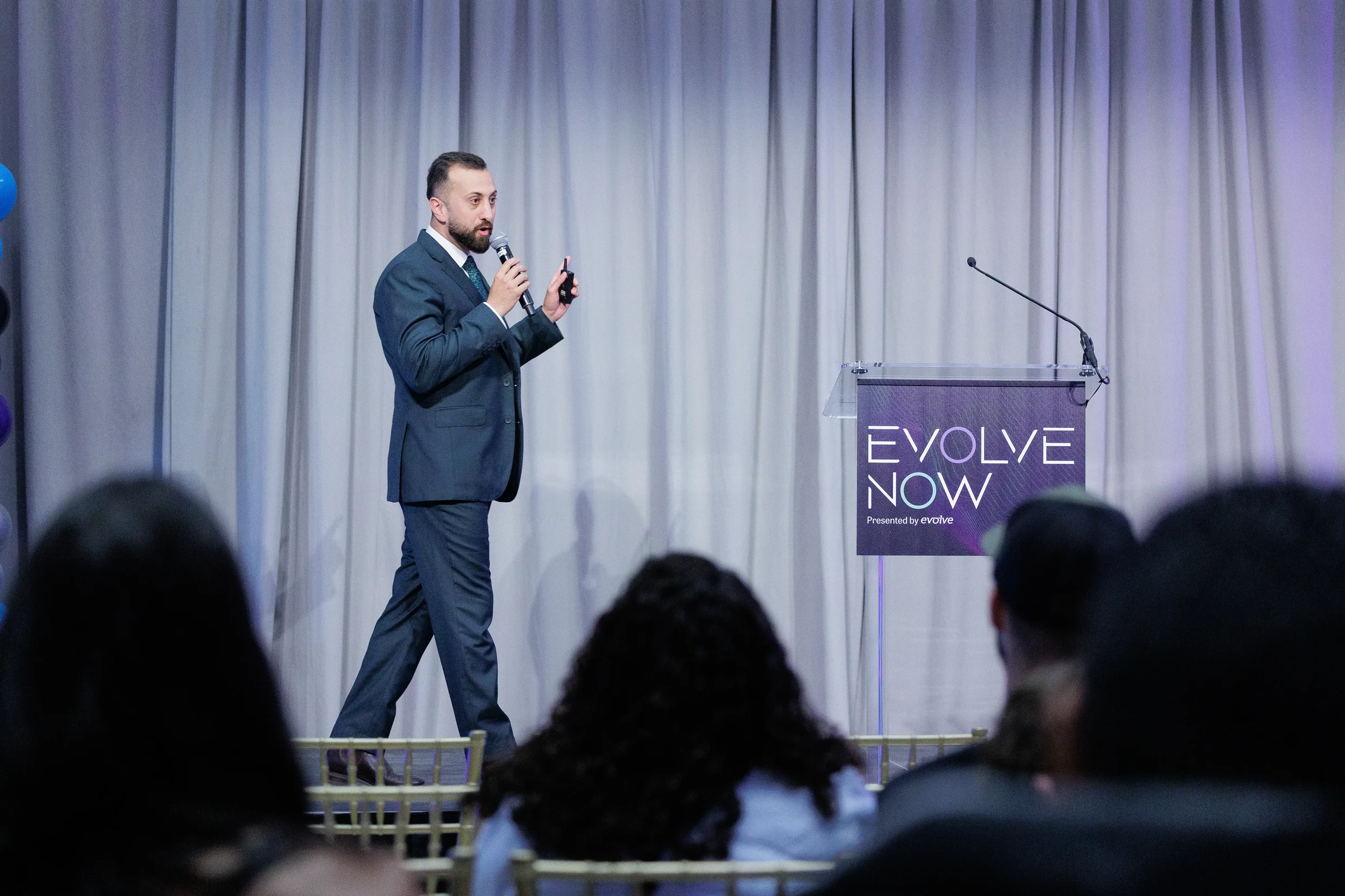Suited speaker walks stage holding microphone with audience silhouette in foreground at Chicago corporate event