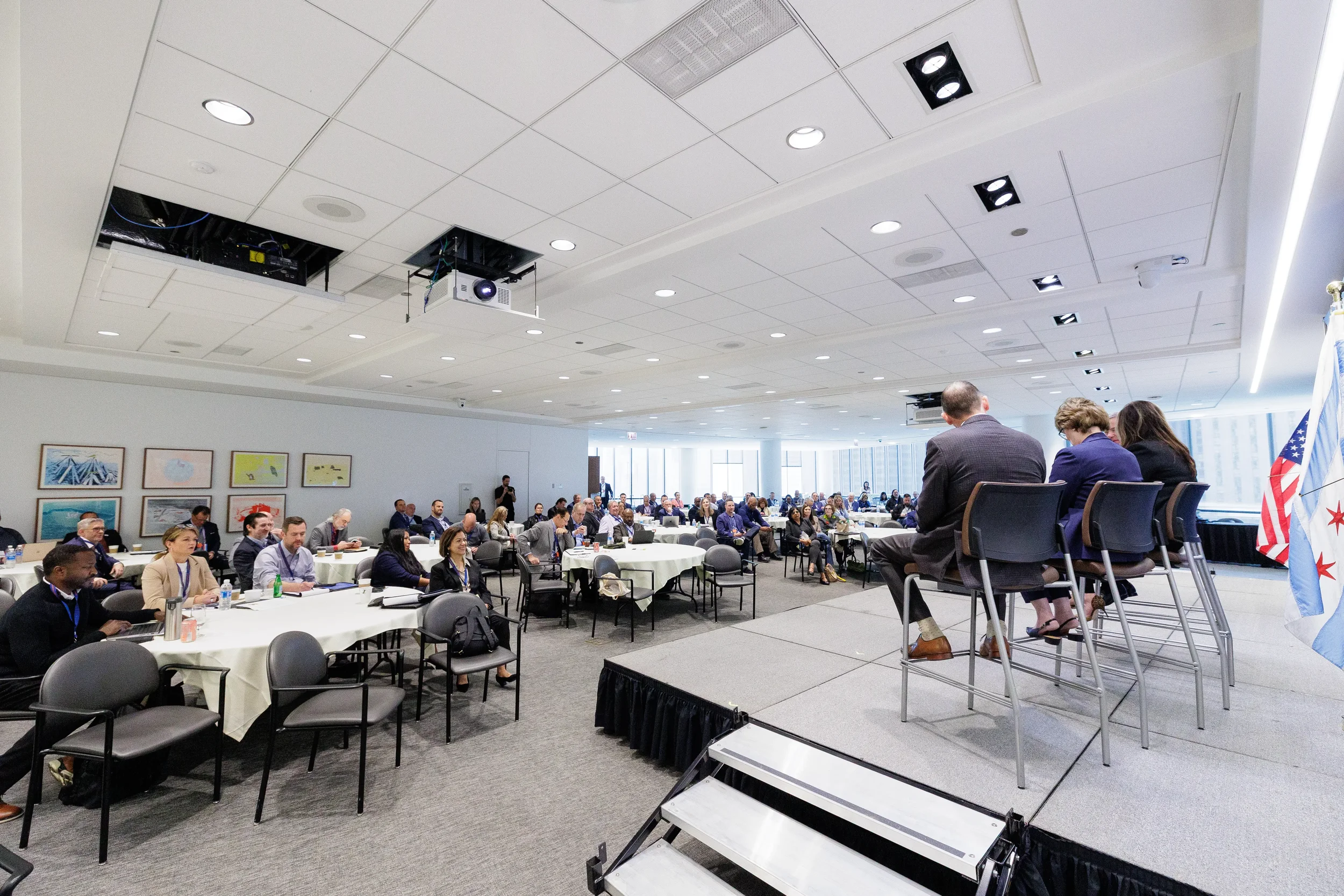 Panelists seated on stools on stage before full room of attendees at round tables at Chicago corporate event