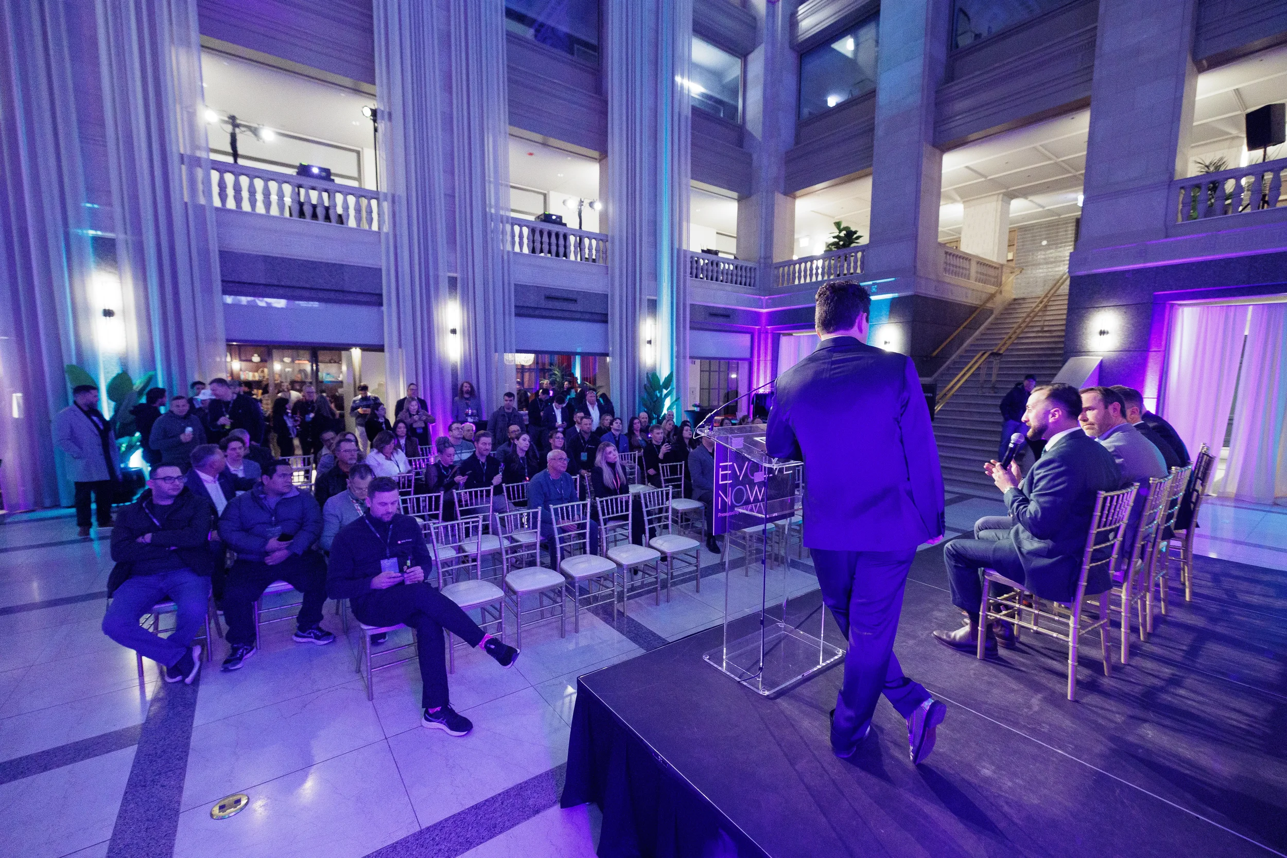 Speaker stands at podium addressing seated audience in grand lit atrium at Chicago corporate event