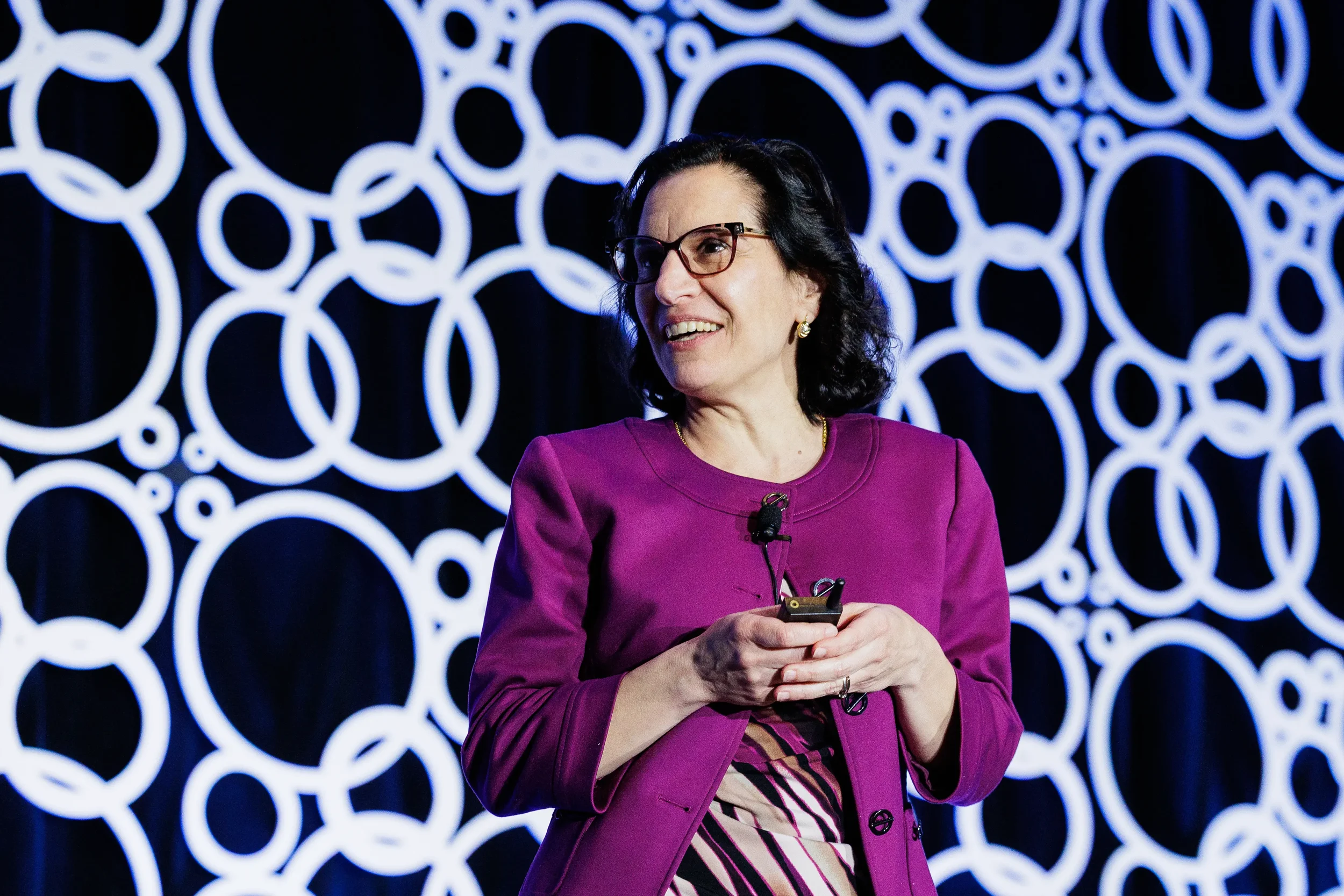 Presenter in magenta jacket smiles holding slide clicker before circle-patterned backdrop at Chicago conference
