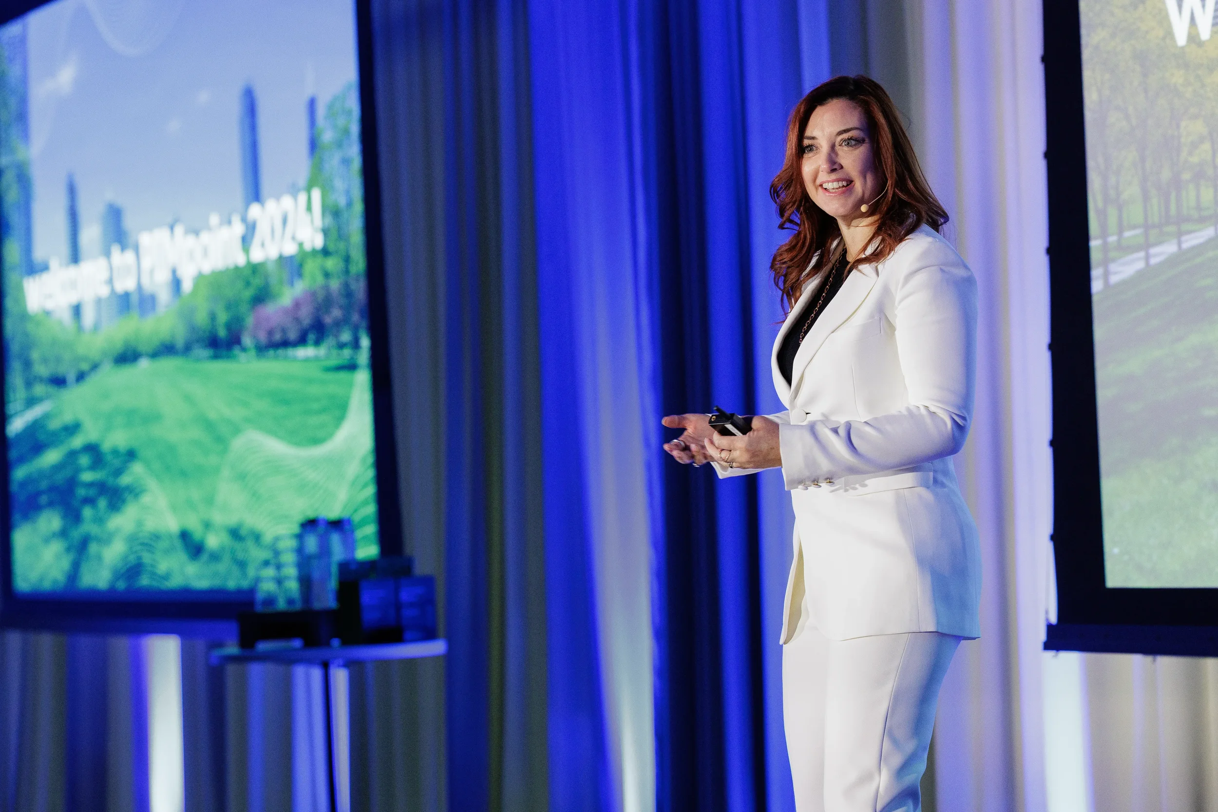 Smiling speaker in white suit presents on stage beside blue curtain at Orlando industry conference