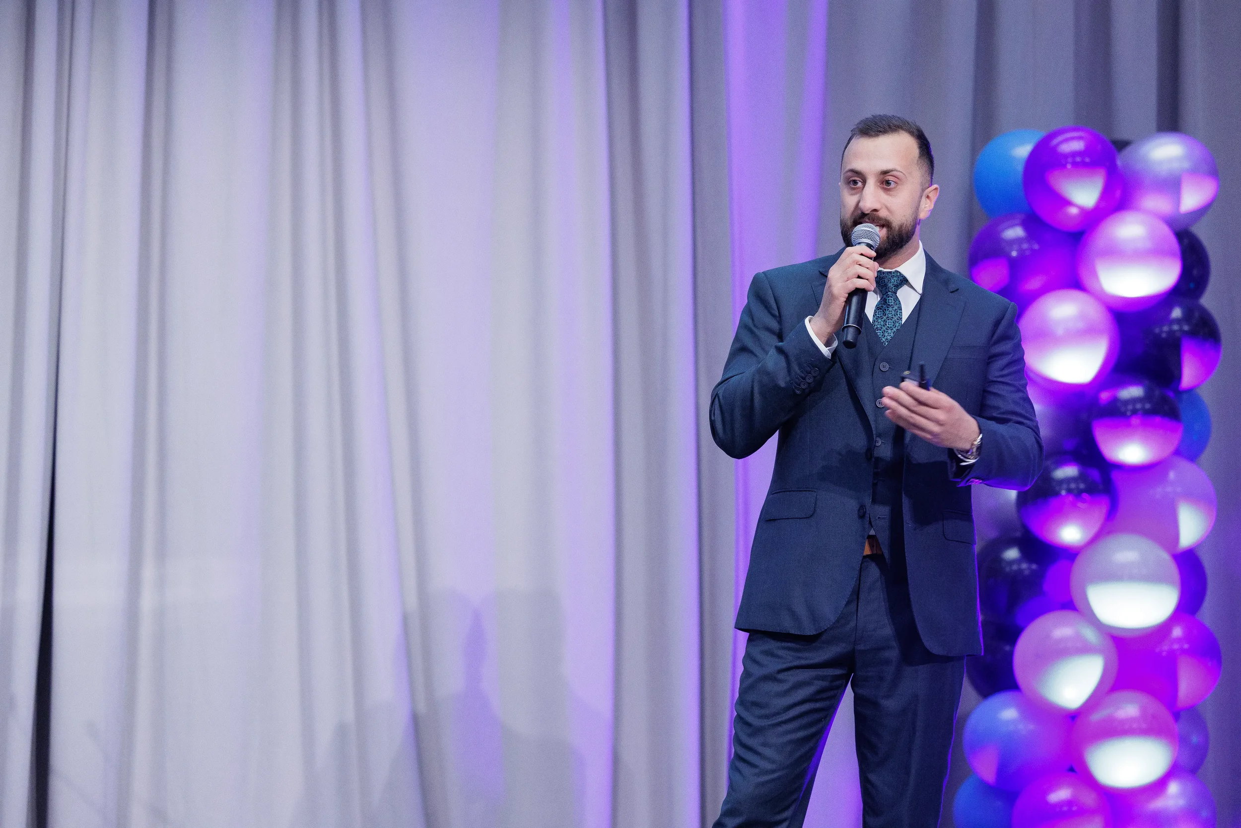 Suited speaker holds microphone on stage beside balloon column at Orlando industry corporate event